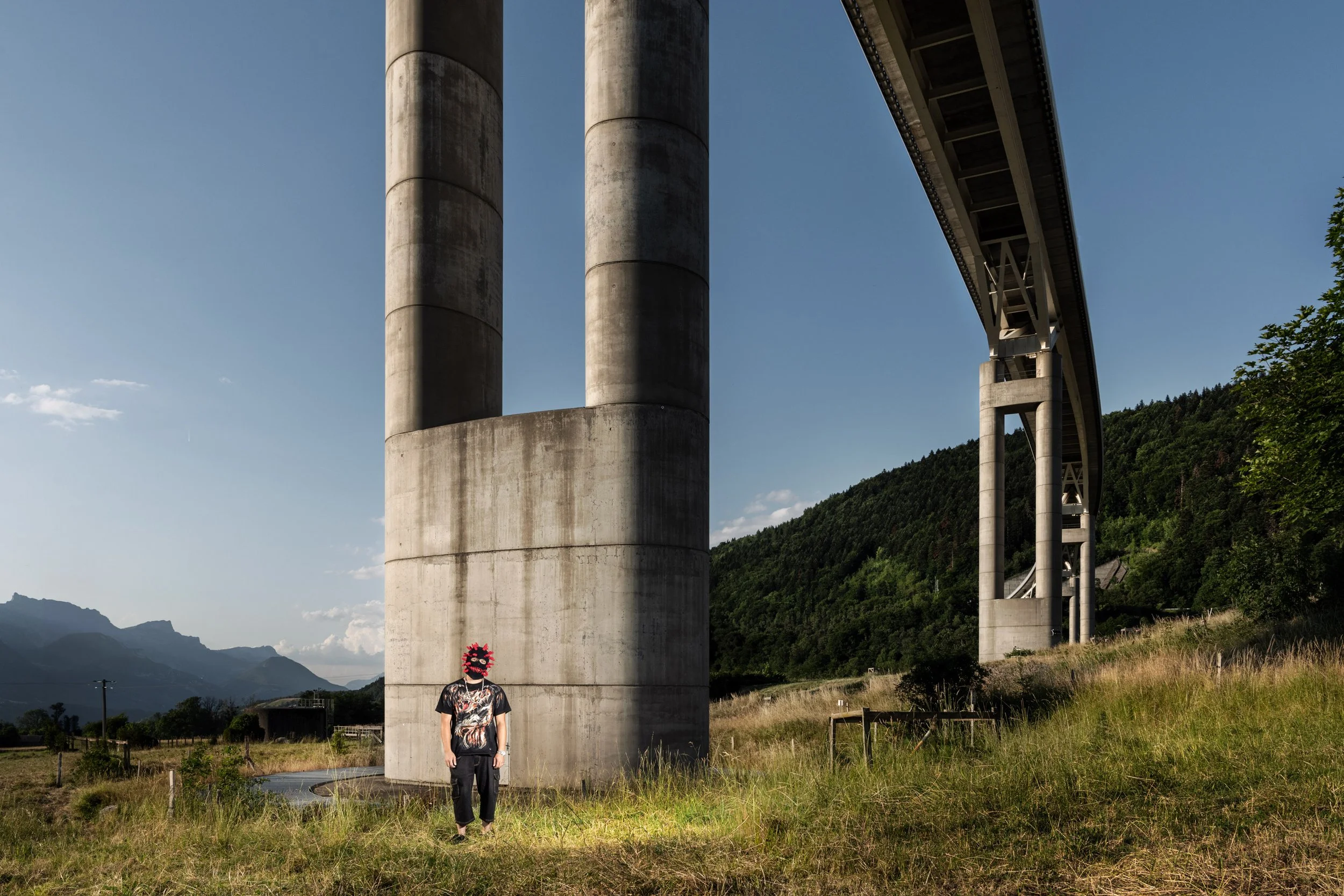 Personne portant un masque rouge et un T-shirt à motifs, debout dans un champ avec un grand pont en béton en arrière-plan.