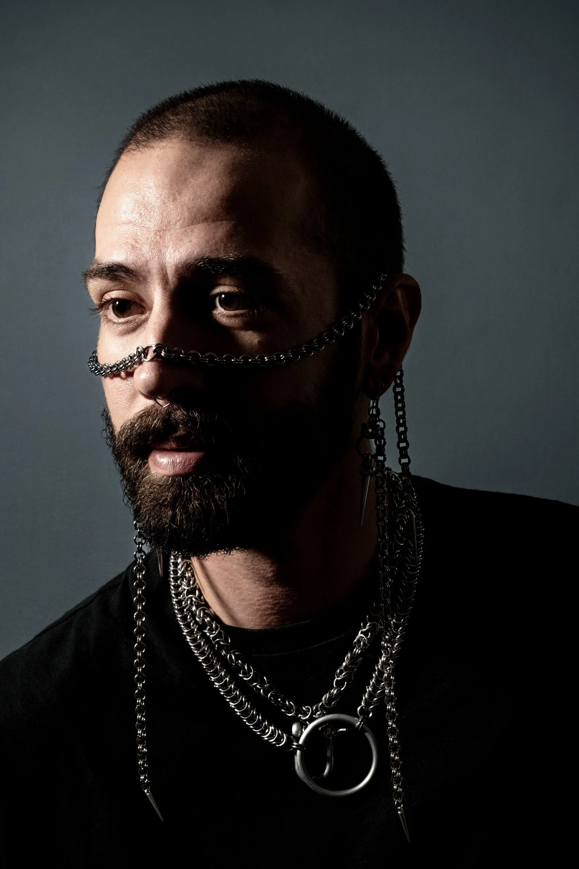 Portrait d'un homme avec des bijoux en chaînes en métal, portant des boucles d'oreilles et un collier massif, avec une expression sérieuse et un éclairage sombre.