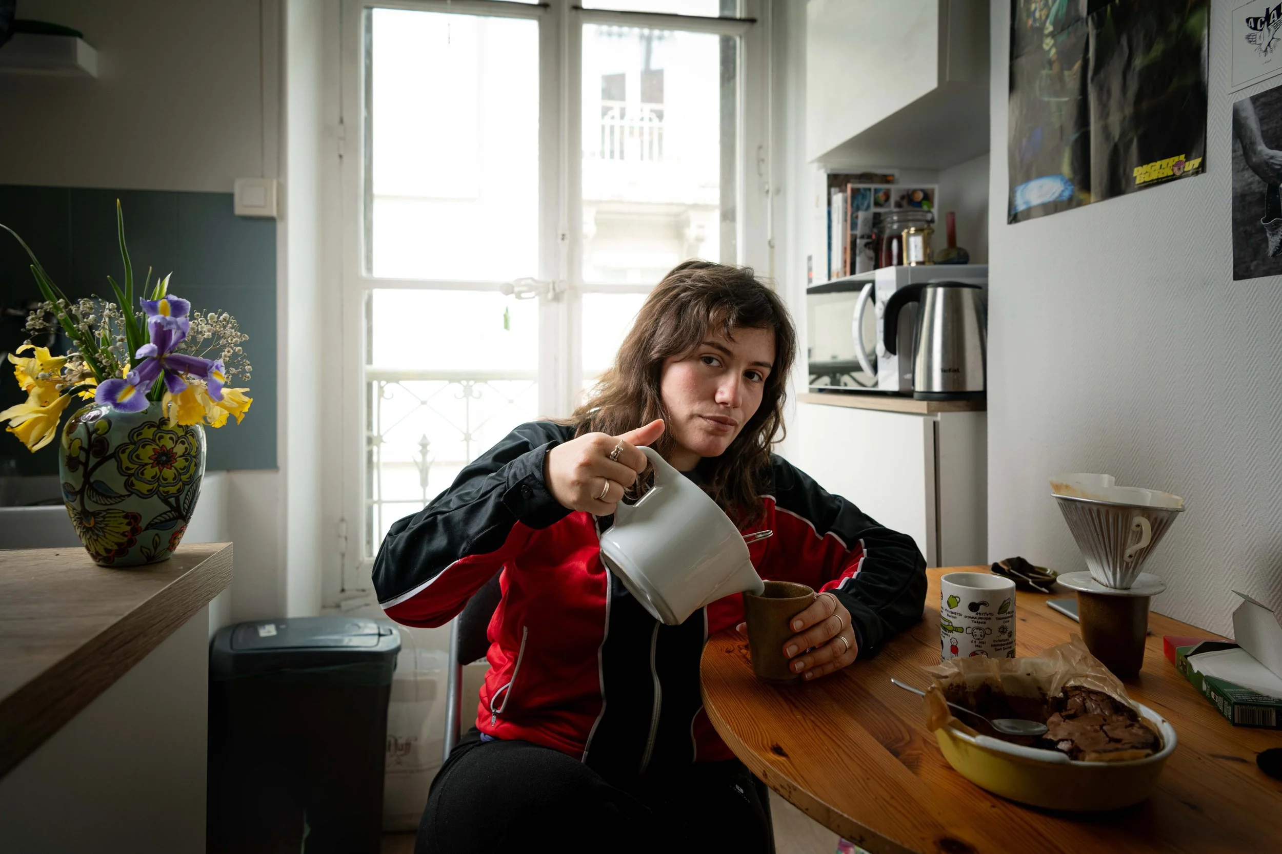 Une jeune femme assise à une table de cuisine, sertant une boisson d'une théière dans une tasse, avec un gâteau au chocolat dans un plat en céramique devant elle, dans une pièce lumineuse avec une porte-fenêtre en arrière-plan, des posters sur le mur