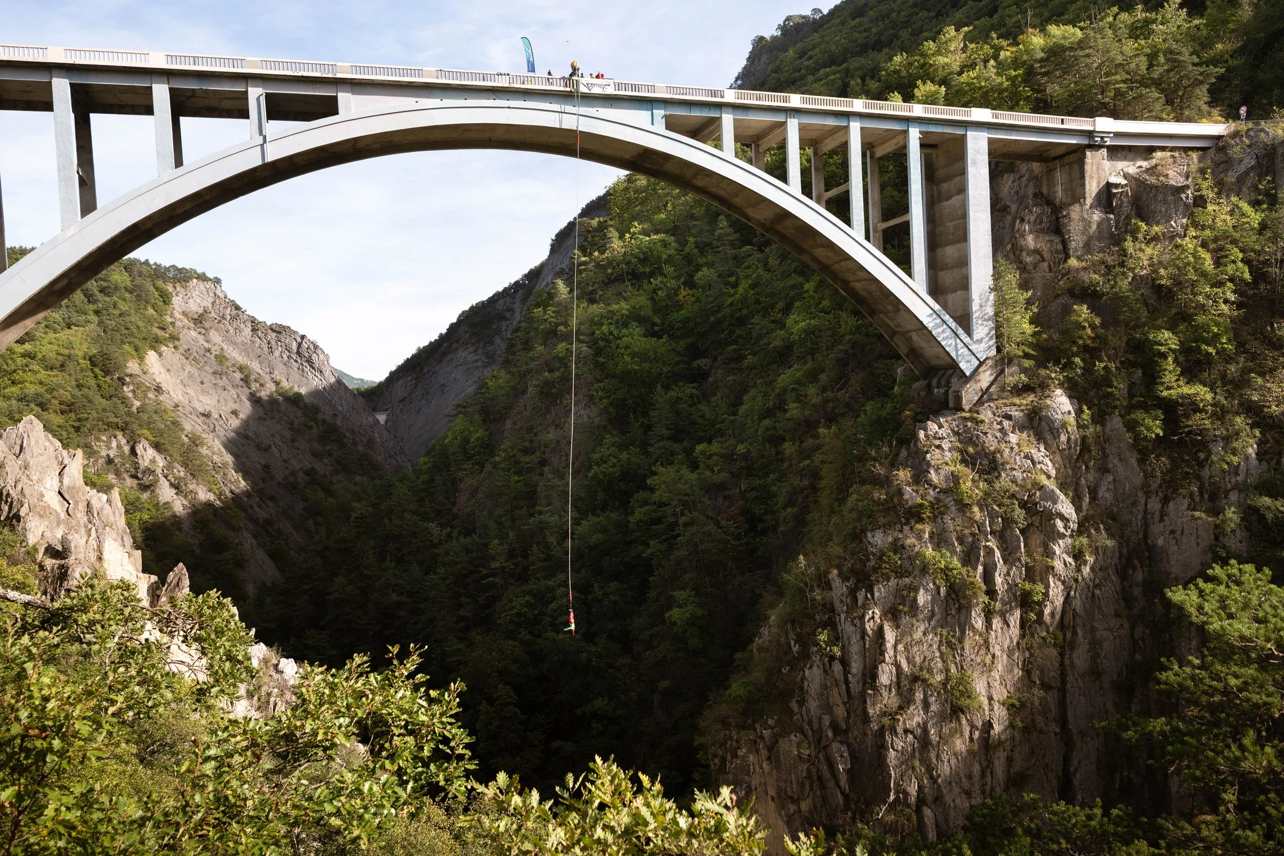 Vue d'une grande arche de pont en béton au-dessus d'une gorge rocheuse couverte de végétation dense, avec un câble de descente suspendu au centre.