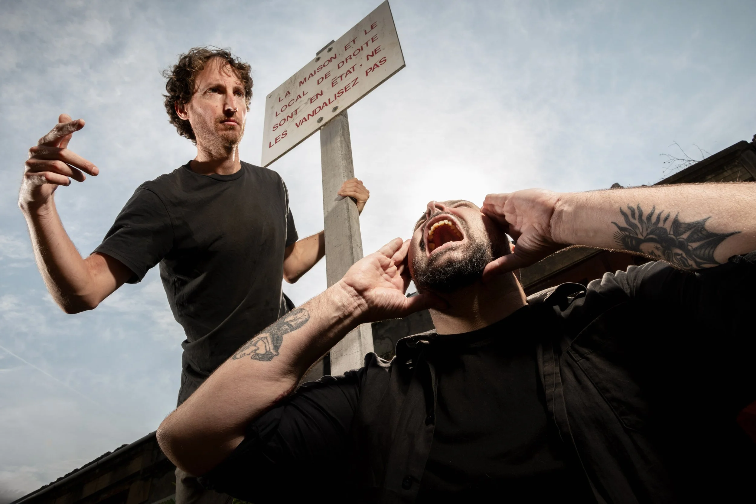 Deux hommes, l'un criant et l'autre tenant une pancarte, semblent engagés dans une dispute ou une confrontation en extérieur, sous un ciel nuageux.