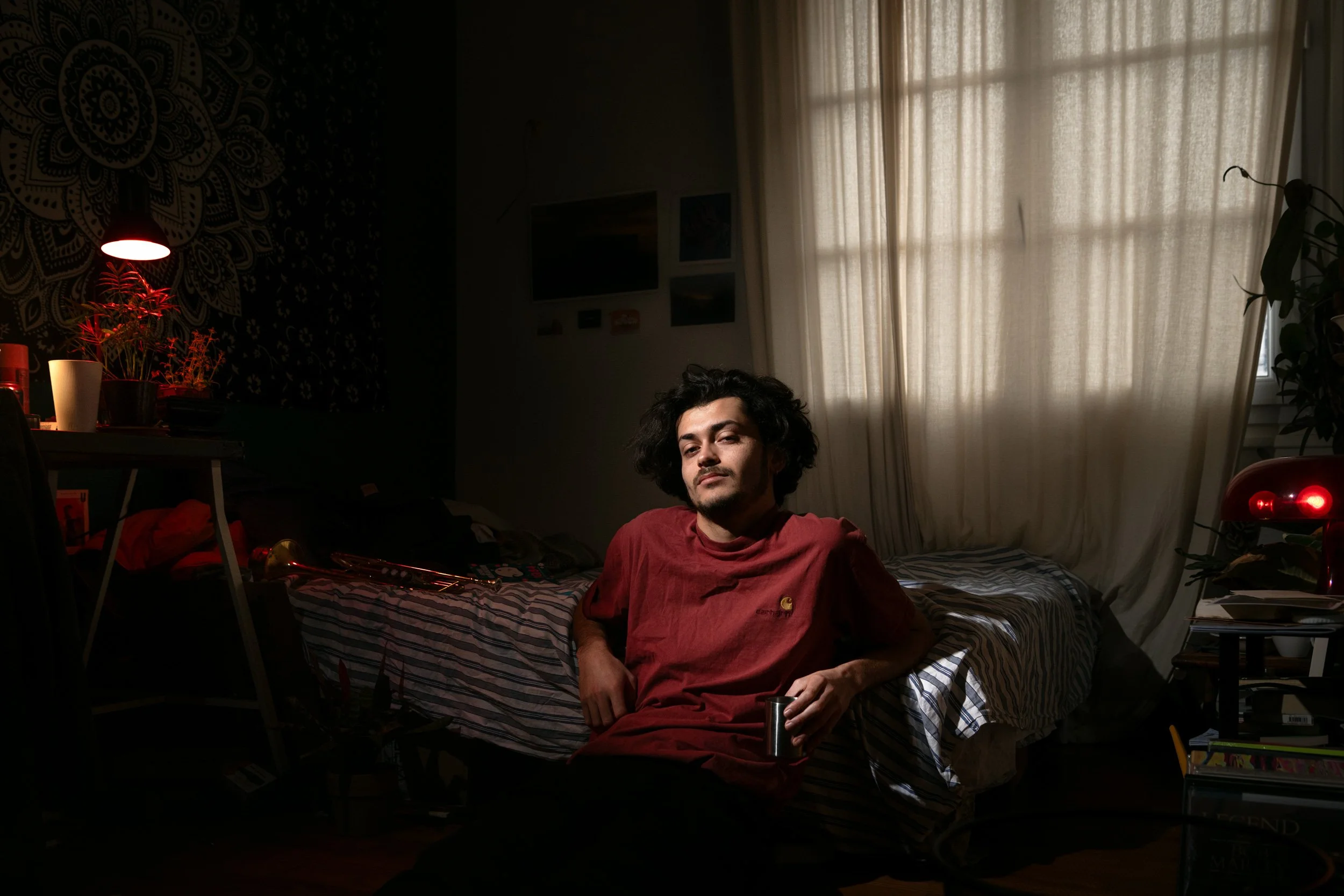 Jeune homme assis sur un lit dans une chambre sombre éclairée par une lampe rouge, avec de la lumière naturelle filtrant à travers des rideaux pâles.