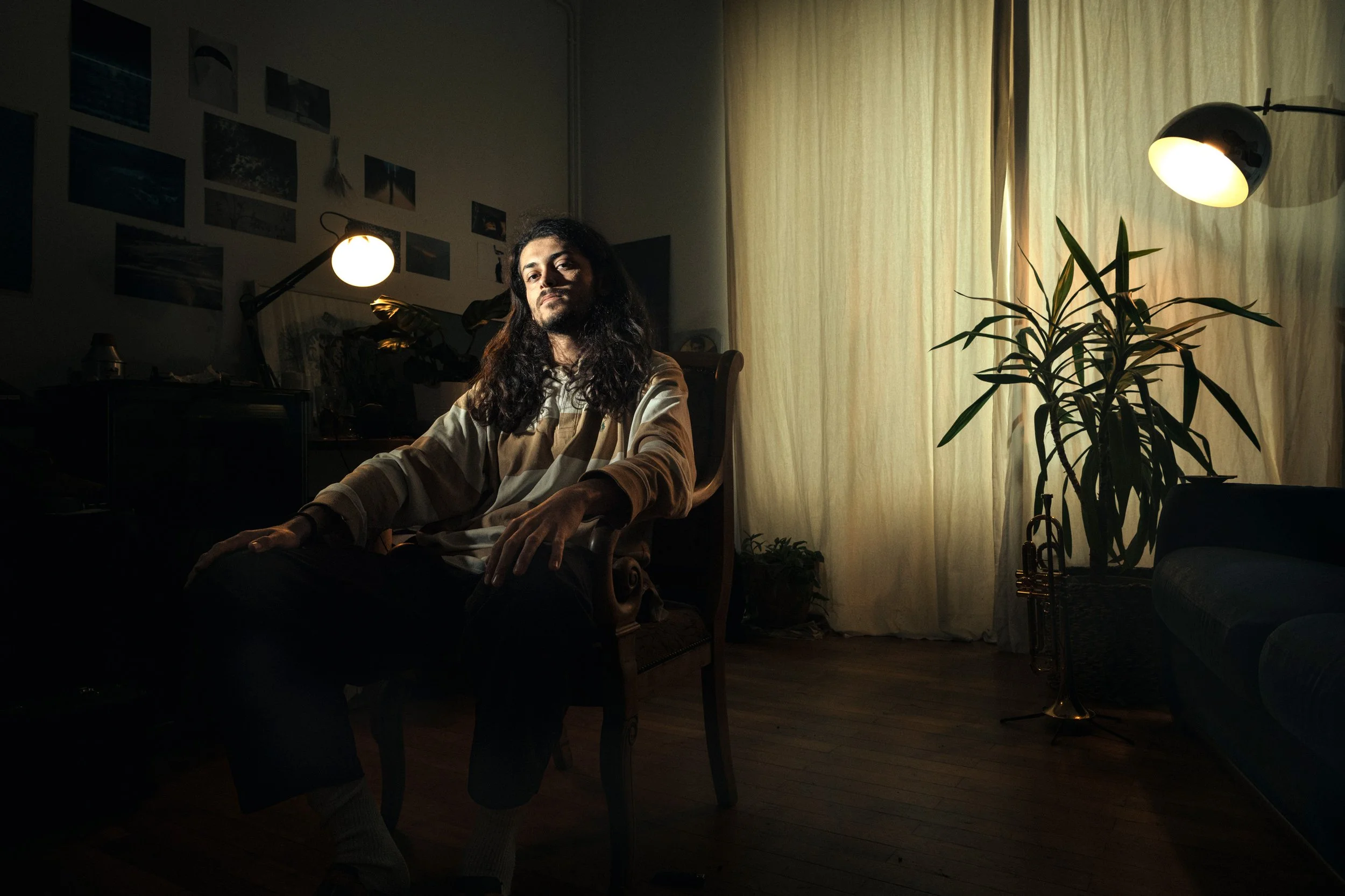 Un homme avec de longs cheveux bouclés assis dans une pièce tamisée, éclairée par deux lampes, avec une grande plante et une fenêtre avec des rideaux jaunes.