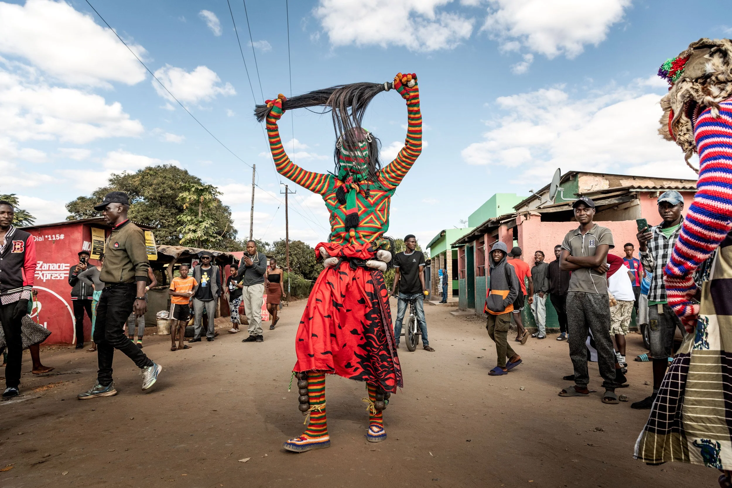 Un danseur africain en costume coloré effectuant une danse dans une rue, entouré d'un groupe de personnes qui regardent et prennent des photos.