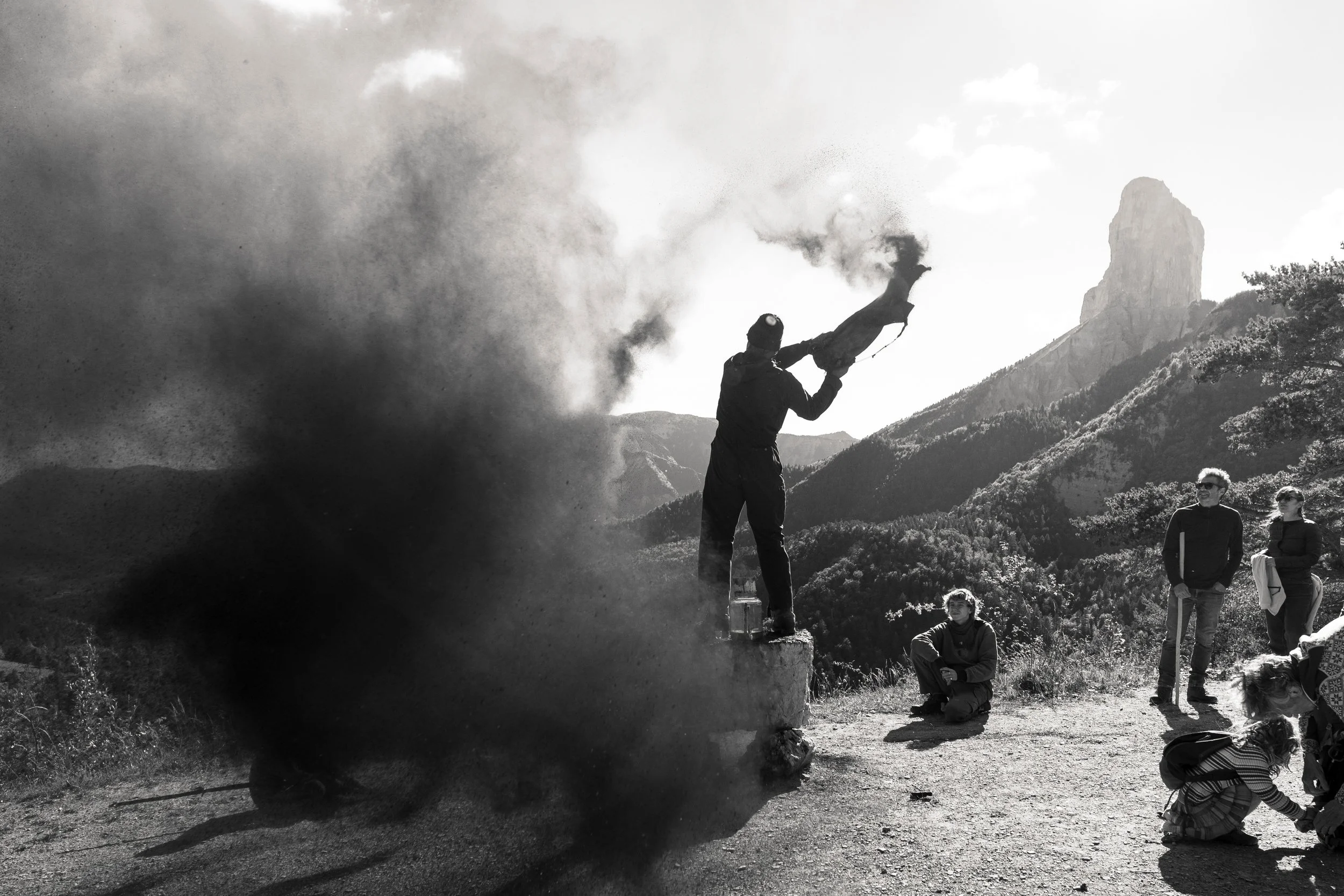 Un groupe de personnes observe un homme qui brûle un objet en montagne, avec un paysage de montagnes en arrière-plan, en noir et blanc. Photographie prise dans le Trièves à l'occasion de la fête des Charbonniers