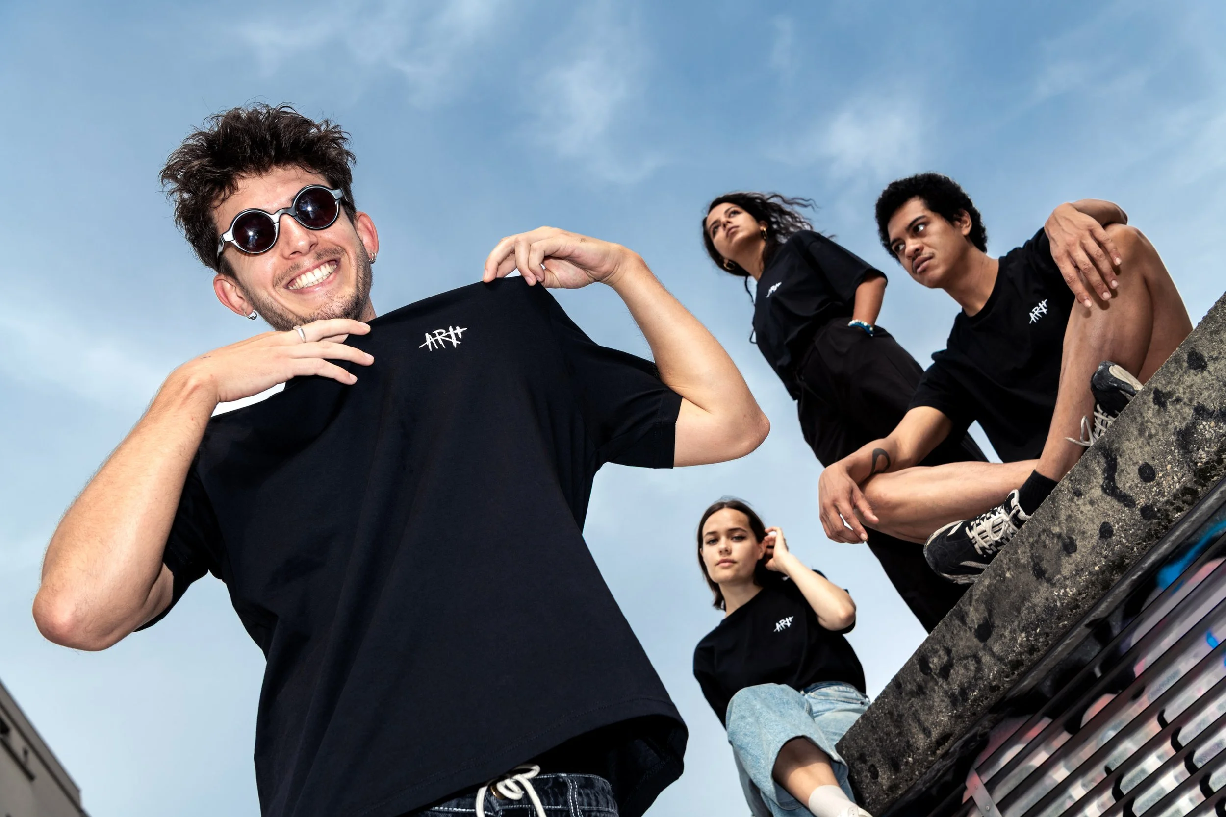 Groupe de jeunes portant des T-shirts noirs avec le logo ARTX, certains assis sur une structure en béton, sous un ciel bleu.