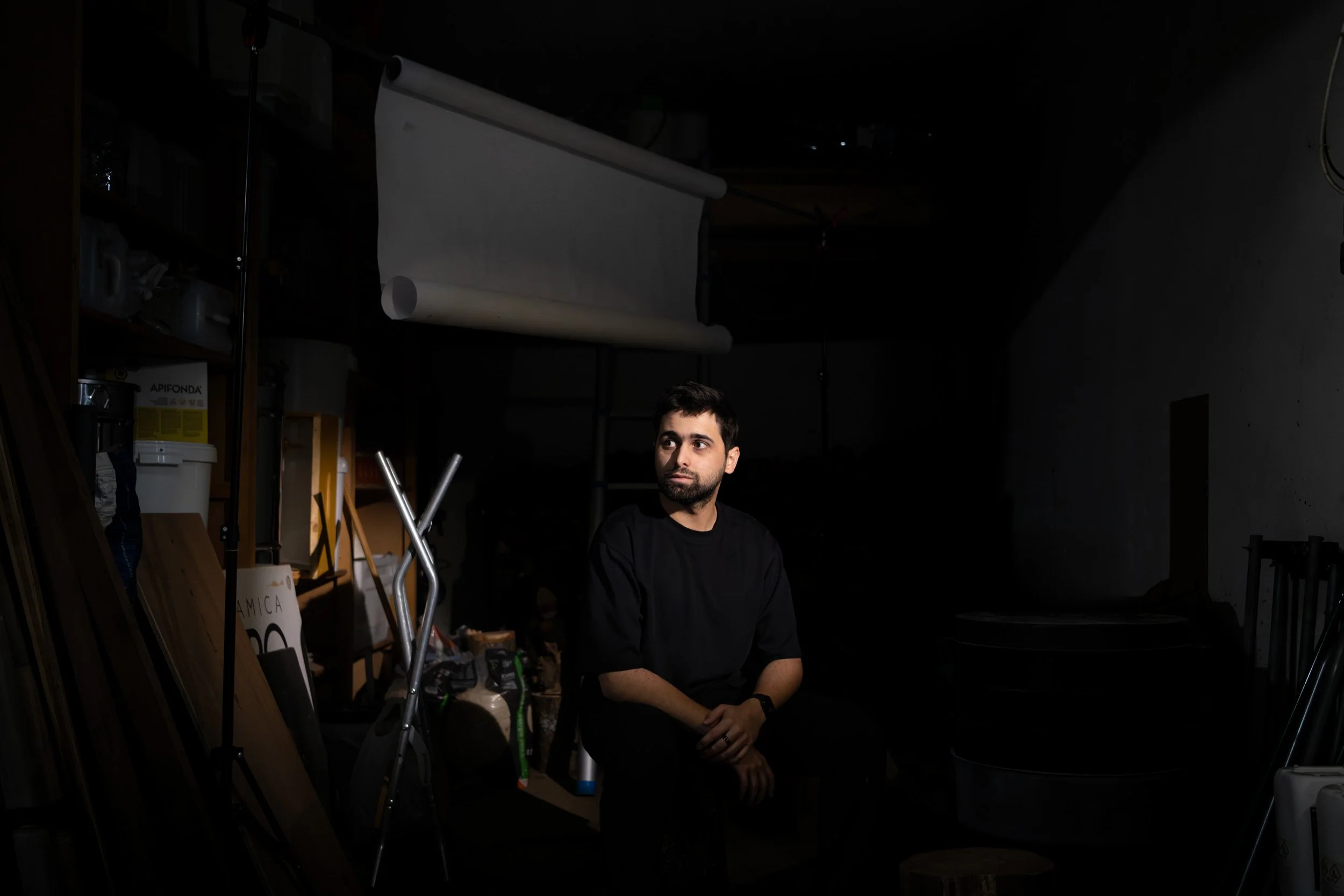 Un homme assis dans une pièce sombre, avec des étagères remplies d'objets et de bocaux sur le côté gauche, un rouleau d'arrière-plan blanc suspendu au plafond derrière lui.