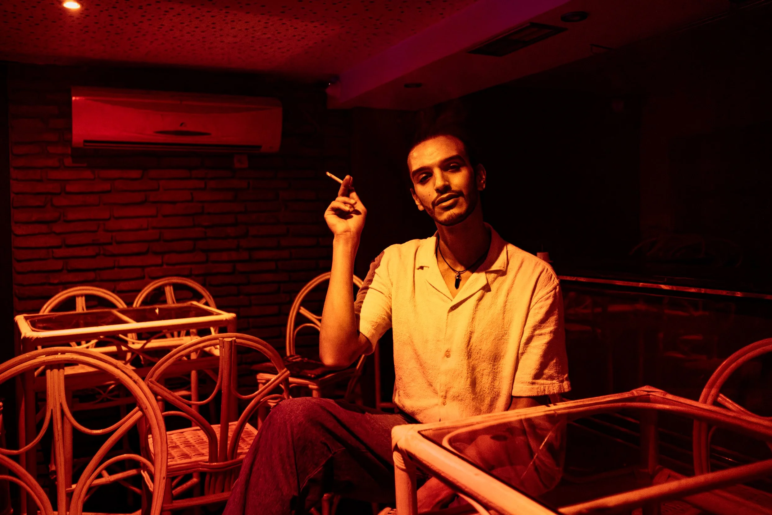 Un homme assis dans un restaurant ou un bar, allumé par une lumière rouge, tenant une cigarette, devant des chaises en rotin, avec un mur en briques en arrière-plan.