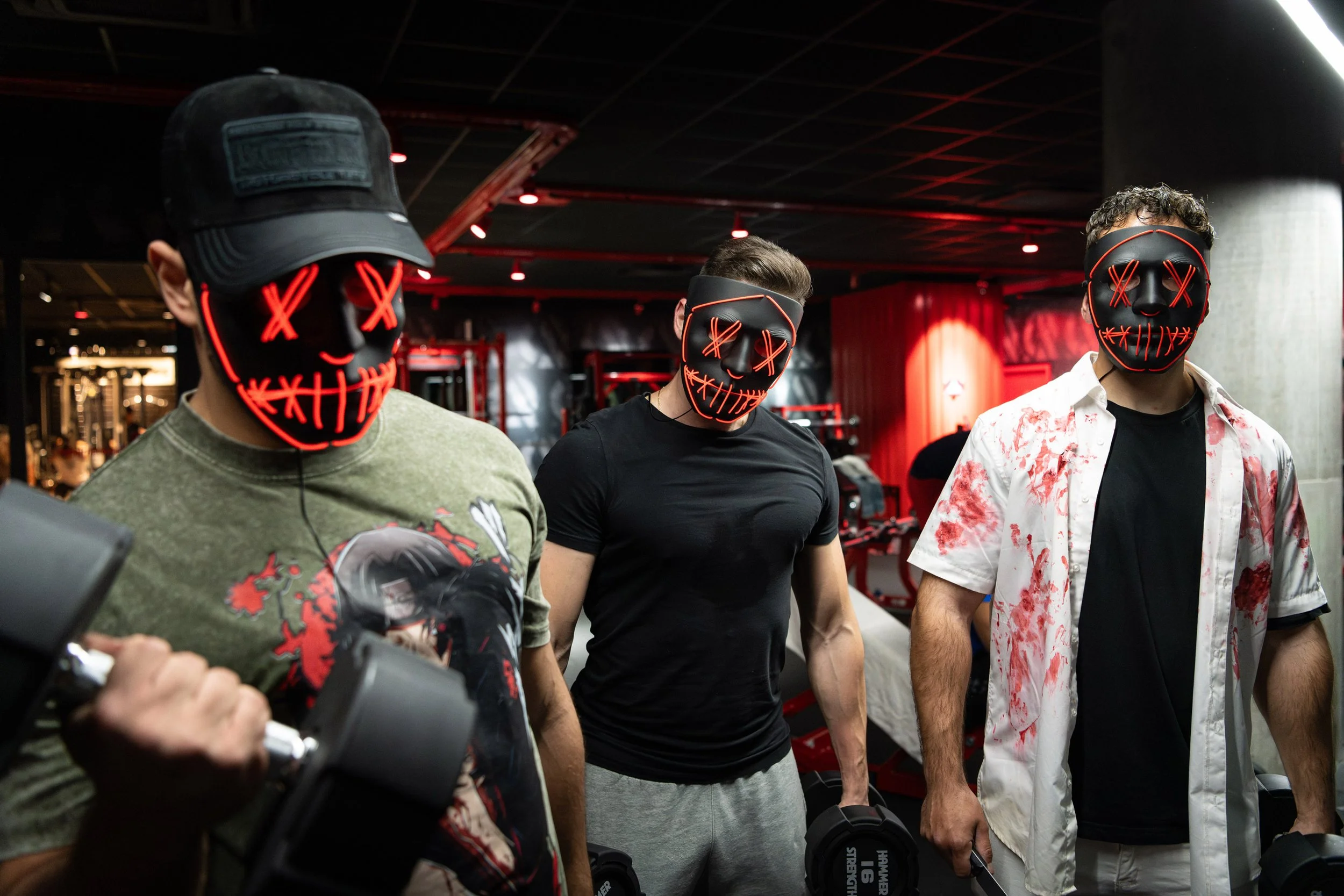 Trois hommes portent des masques noirs avec des motifs de visage souriant lumineux en rouge, dans une salle de sport.