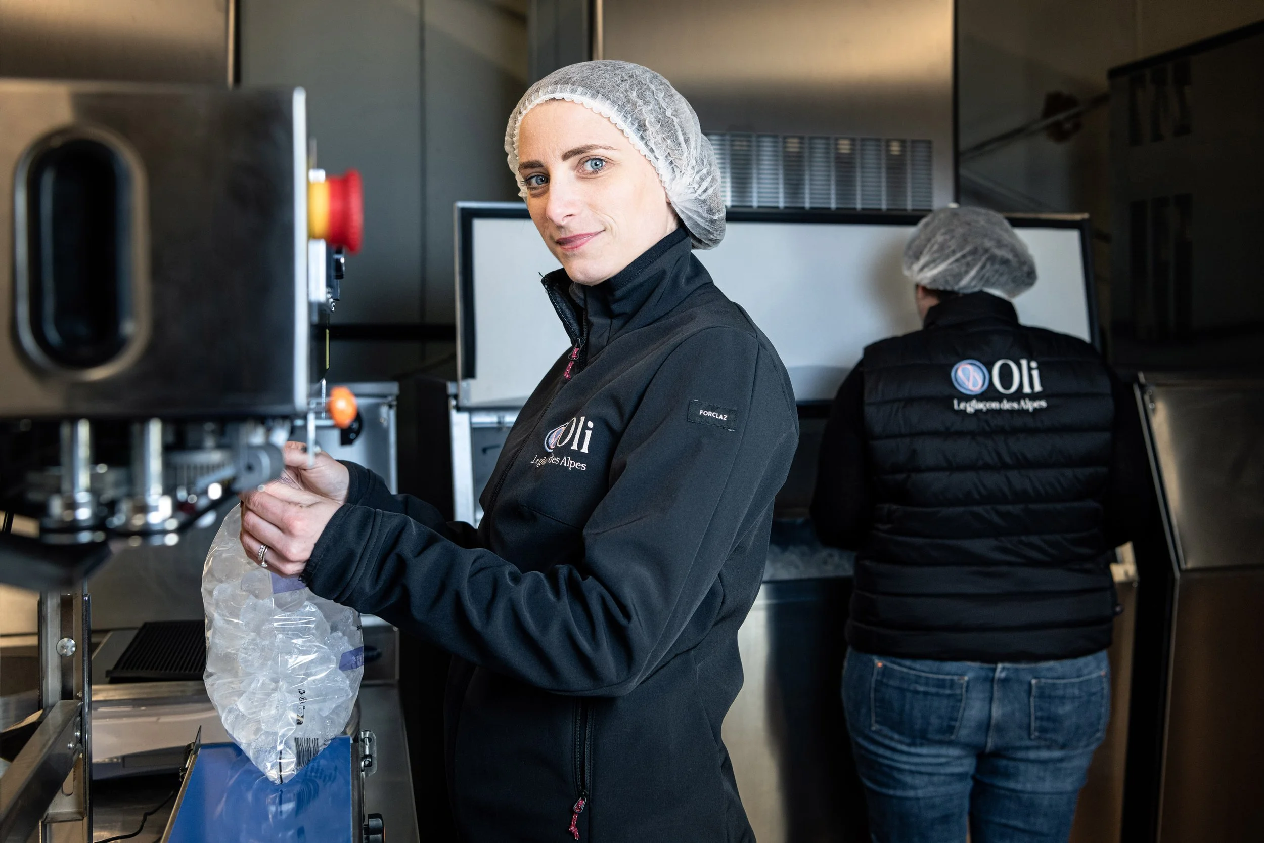 Deux femmes en vêtements de travail, portant des gilets et cagoules, travaillant dans une cuisine ou un laboratoire industriel, avec un logo 'Oli' sur leurs vêtements.