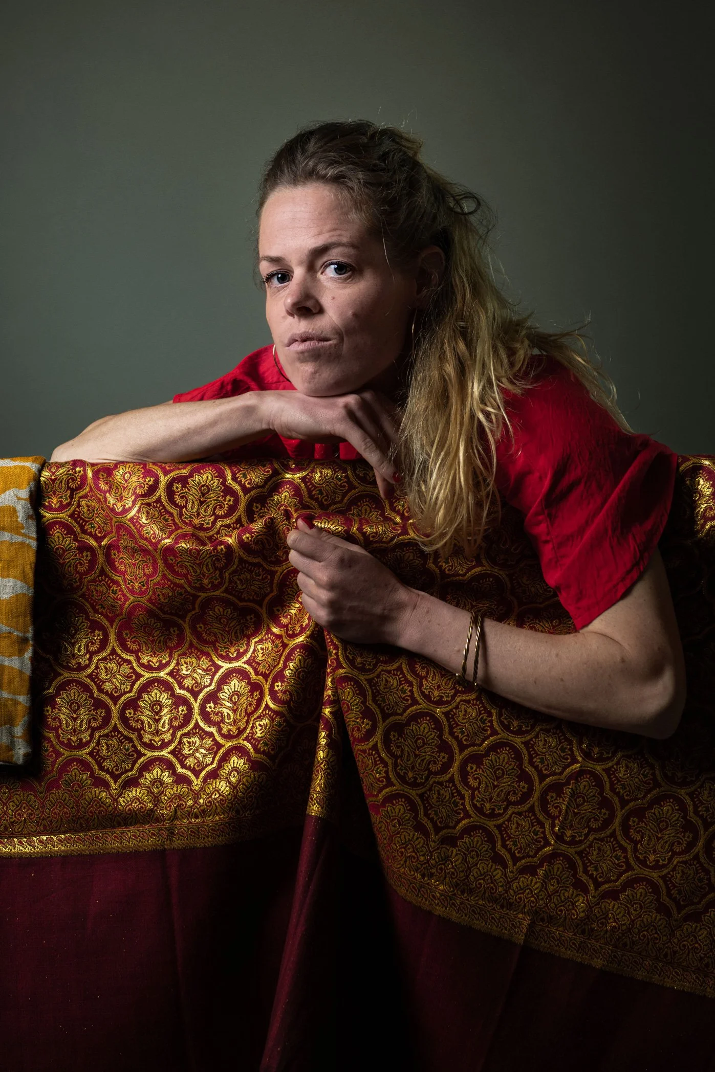 Portrait d'une femme avec des cheveux blonds, portant un t-shirt rouge, posant avec un bras appuyé sur un meuble orné d'un tissu à motifs dorés et rouges, avec un fond gris