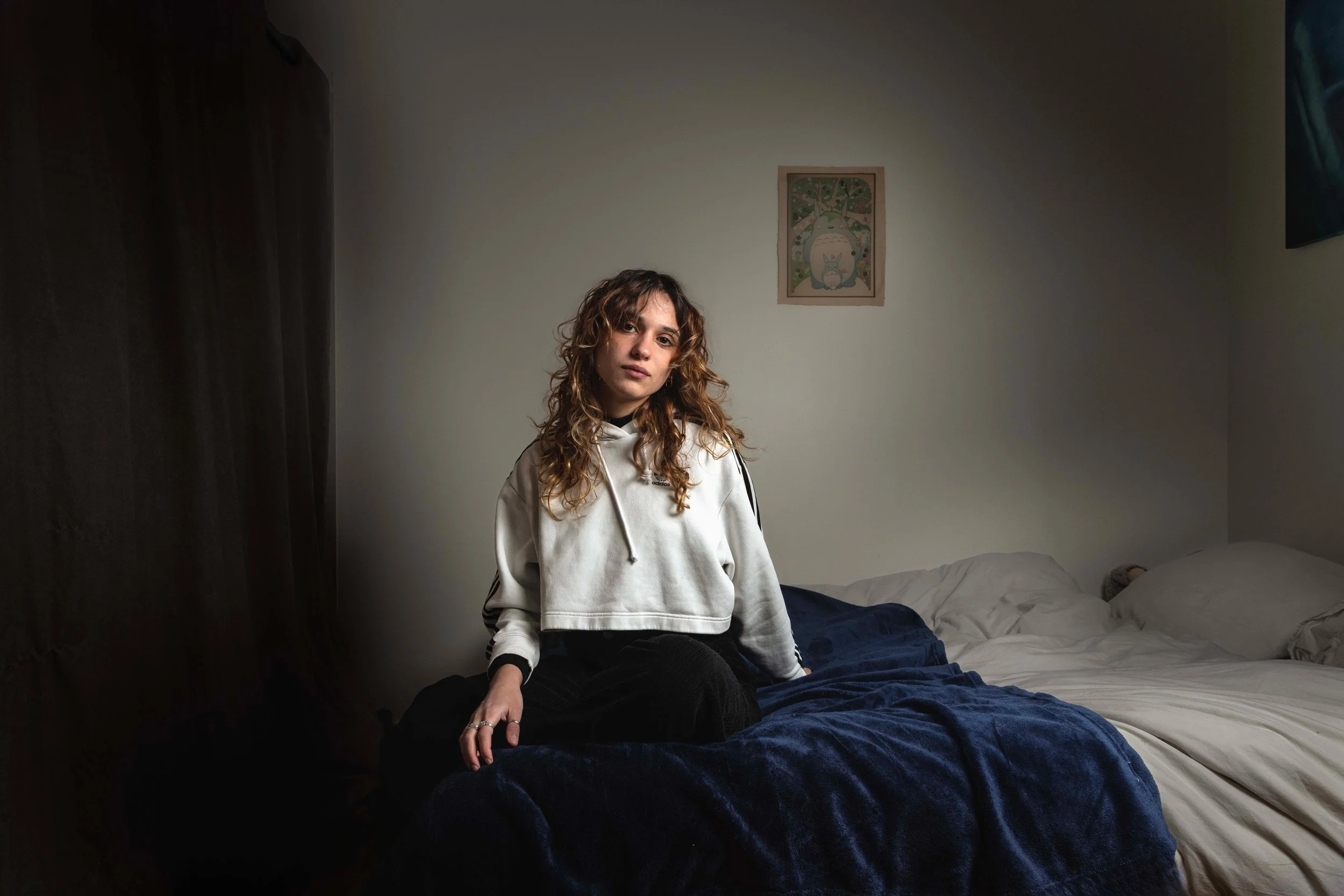 Jeune femme avec des cheveux bouclés assise sur un lit avec une couverture bleue, portant un sweat blanc, dans une chambre avec une peinture murale en arrière-plan.