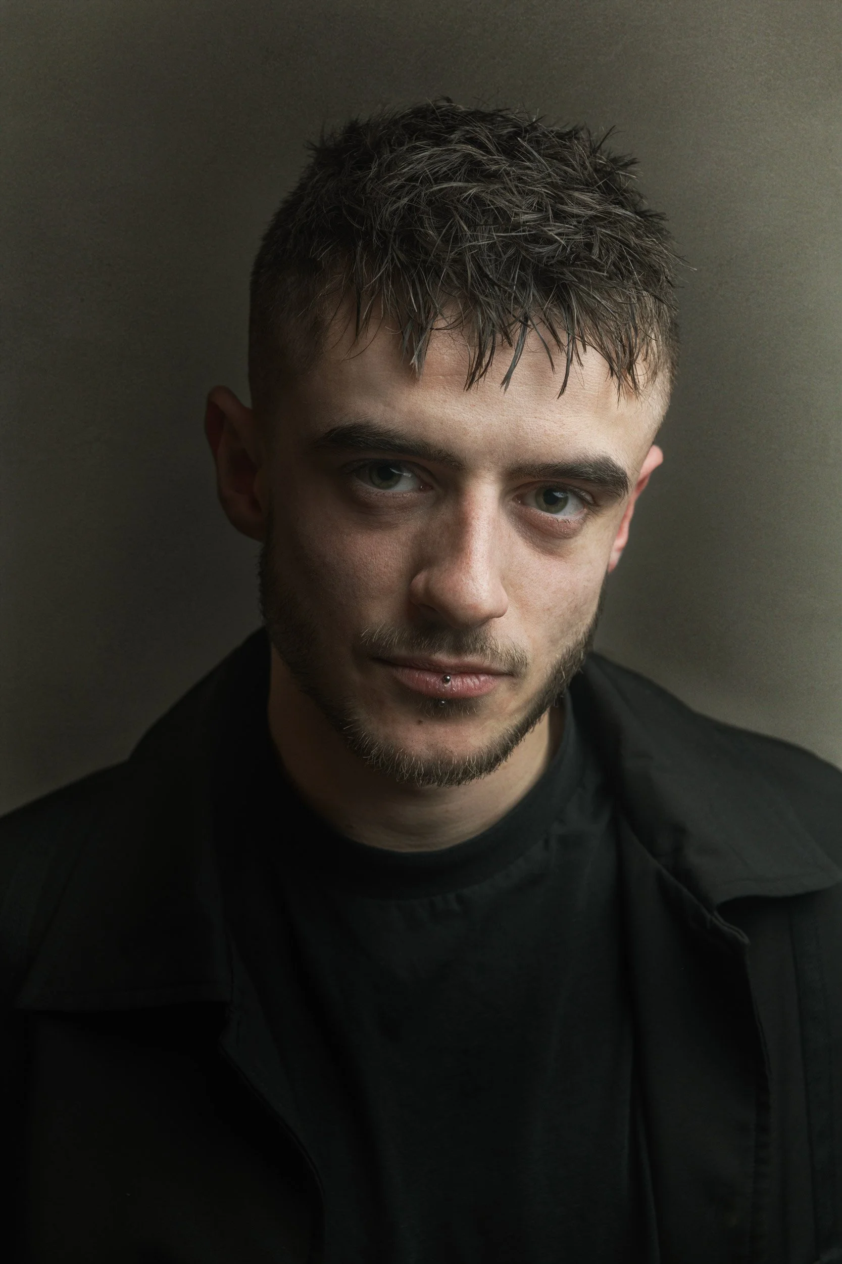 Un jeune homme avec des cheveux courts, une barbe légère, portant un t-shirt noir et un blouson noir, regarde vers l'appareil photo contre un fond neutre.