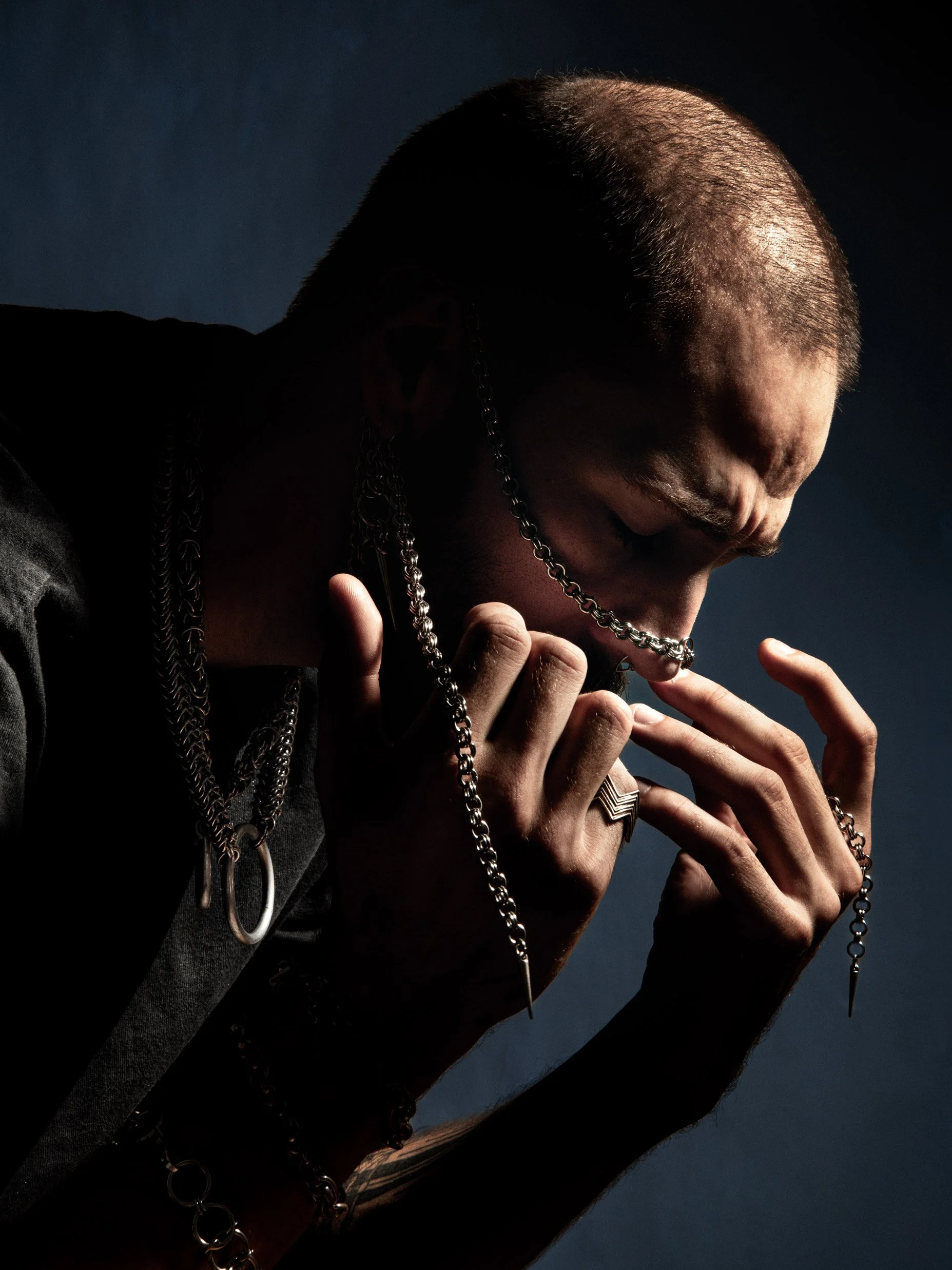 Un homme avec une tête rasée ou chauve, portant plusieurs chaînes en métal, avec une main près de son visage, dans un éclairage sombre.
