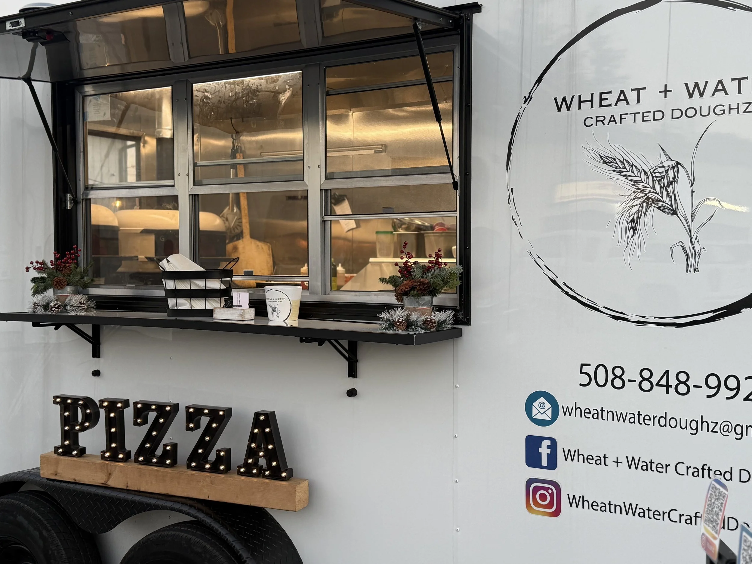 A food truck decorated with holiday plants, featuring a sign that spells out PIZZA with marquee lights. The truck advertises wheat and water crafted dough, with contact info and social media handles displayed.
