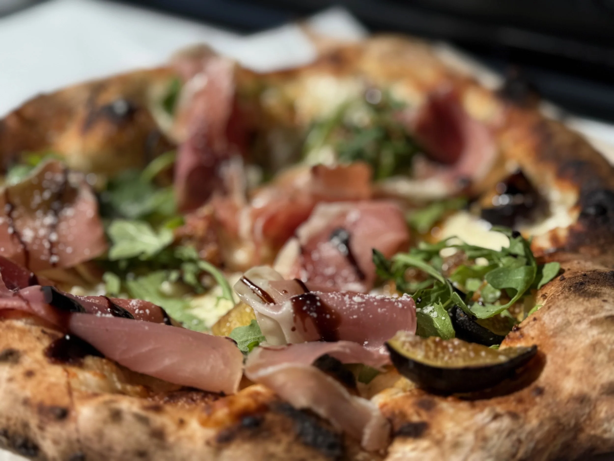 Close-up of a pizza topped with arugula, black olives, prosciutto, and cheese, with a slightly charred crust.