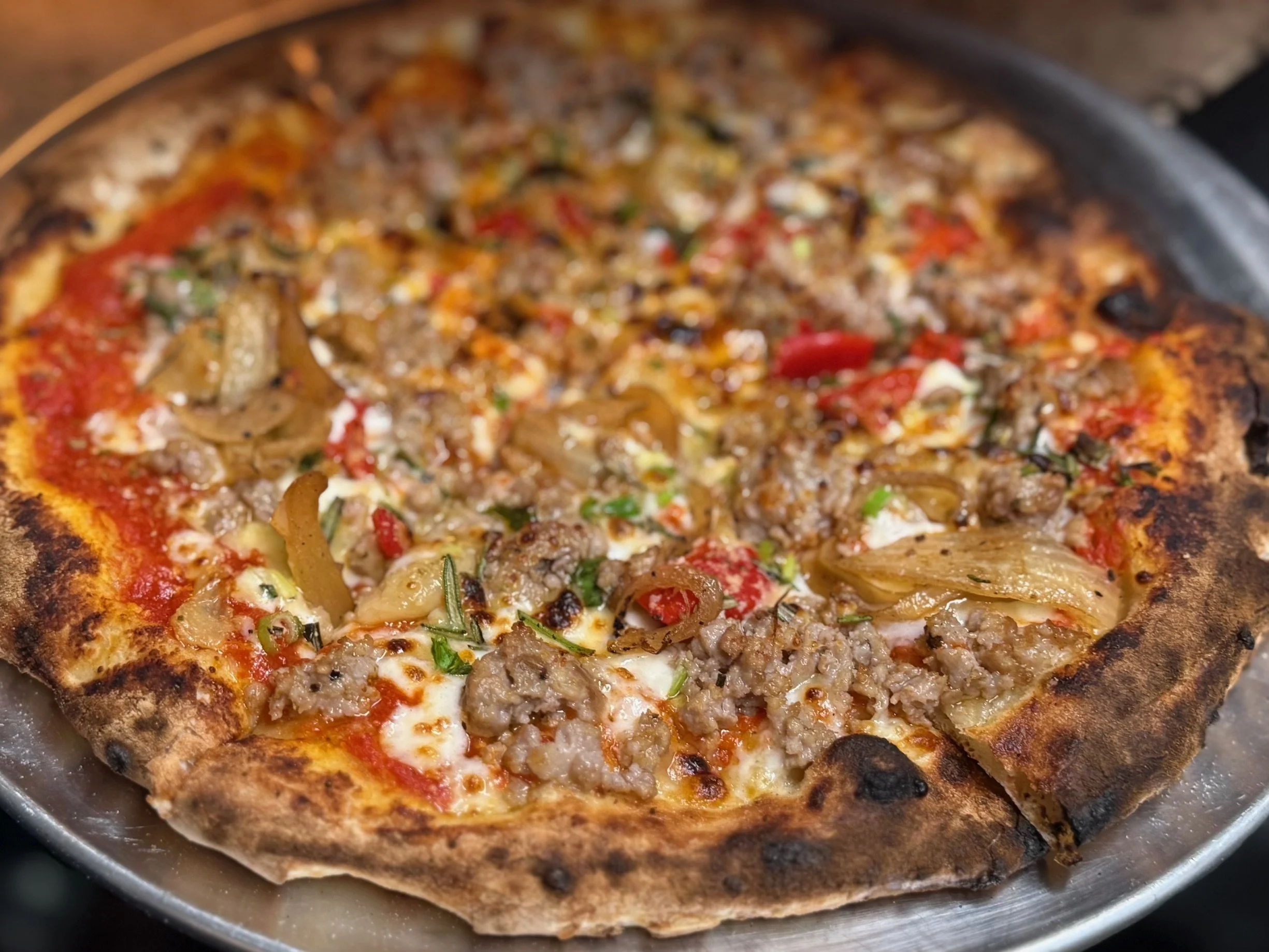 Close-up of a cooked pizza with meat, onions, red peppers, cheese, and tomato sauce on a metal tray.