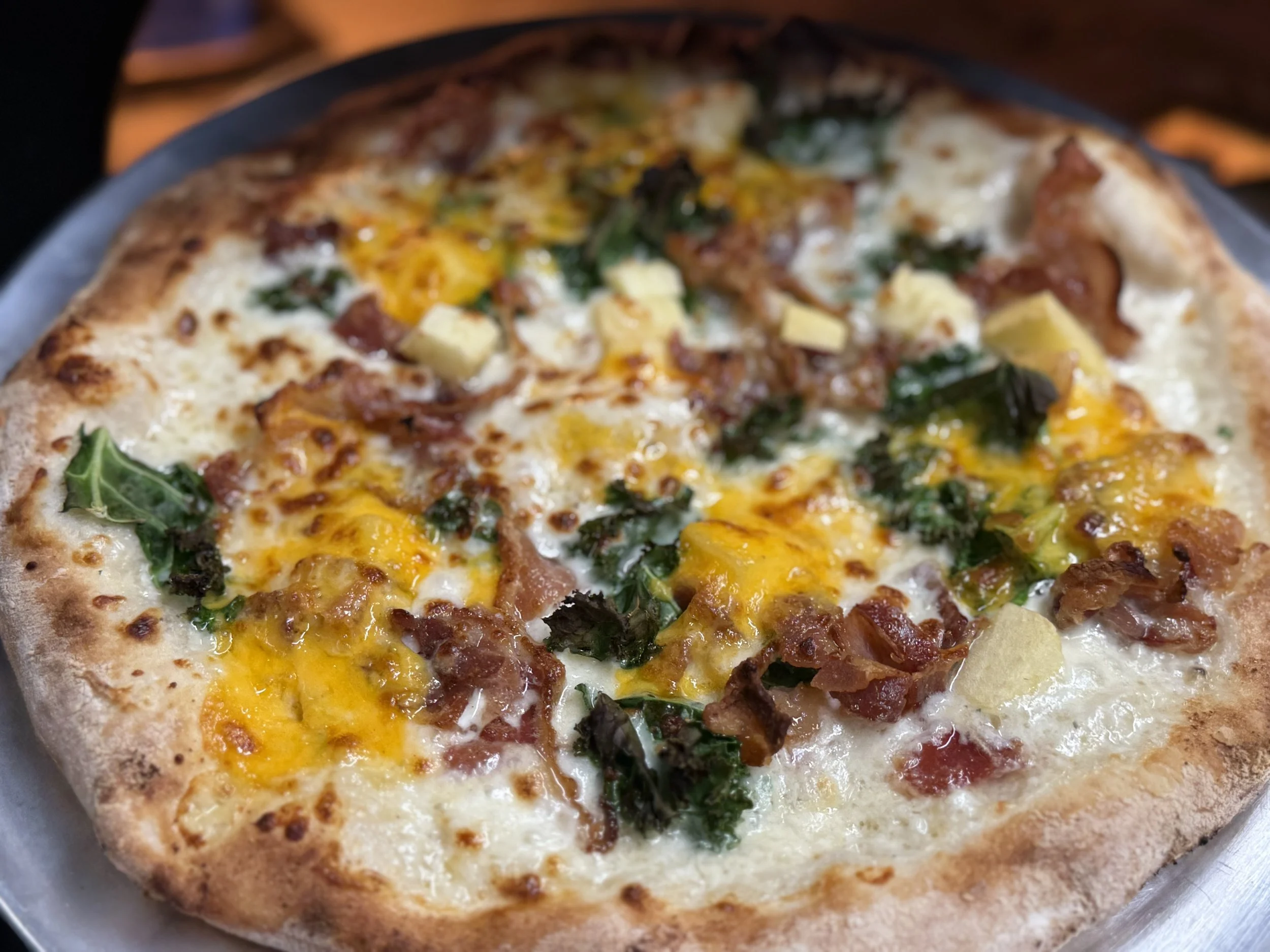 Close-up of a pizza topped with bacon, yellow cheese, green vegetables, and chunks of yellow potato, on a metal tray.