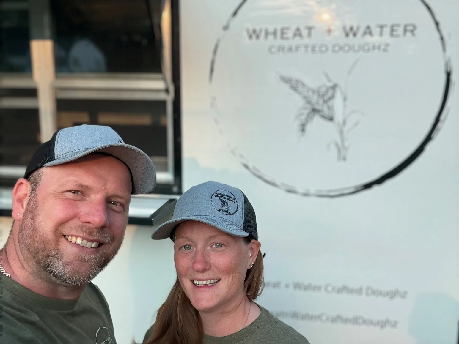 A smiling man and woman taking a selfie in front of a food truck with a sign that reads 'Wheat & Water Crafted Doughz' and features a hummingbird illustration.
