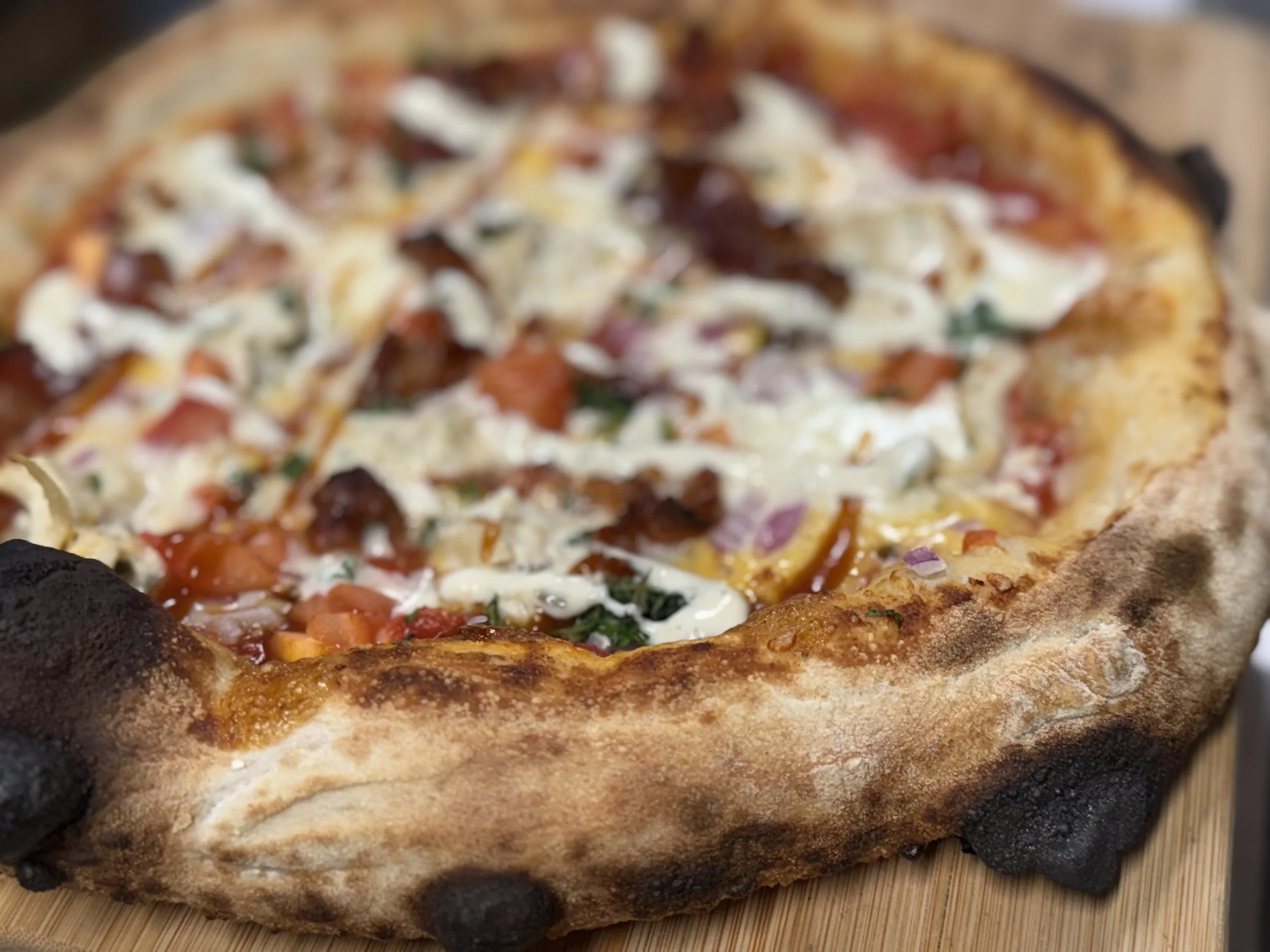Close-up of a cooked pizza with a thick crust and burnt edges, topped with cheese, tomato sauce, vegetables, and meat, on a wooden surface.