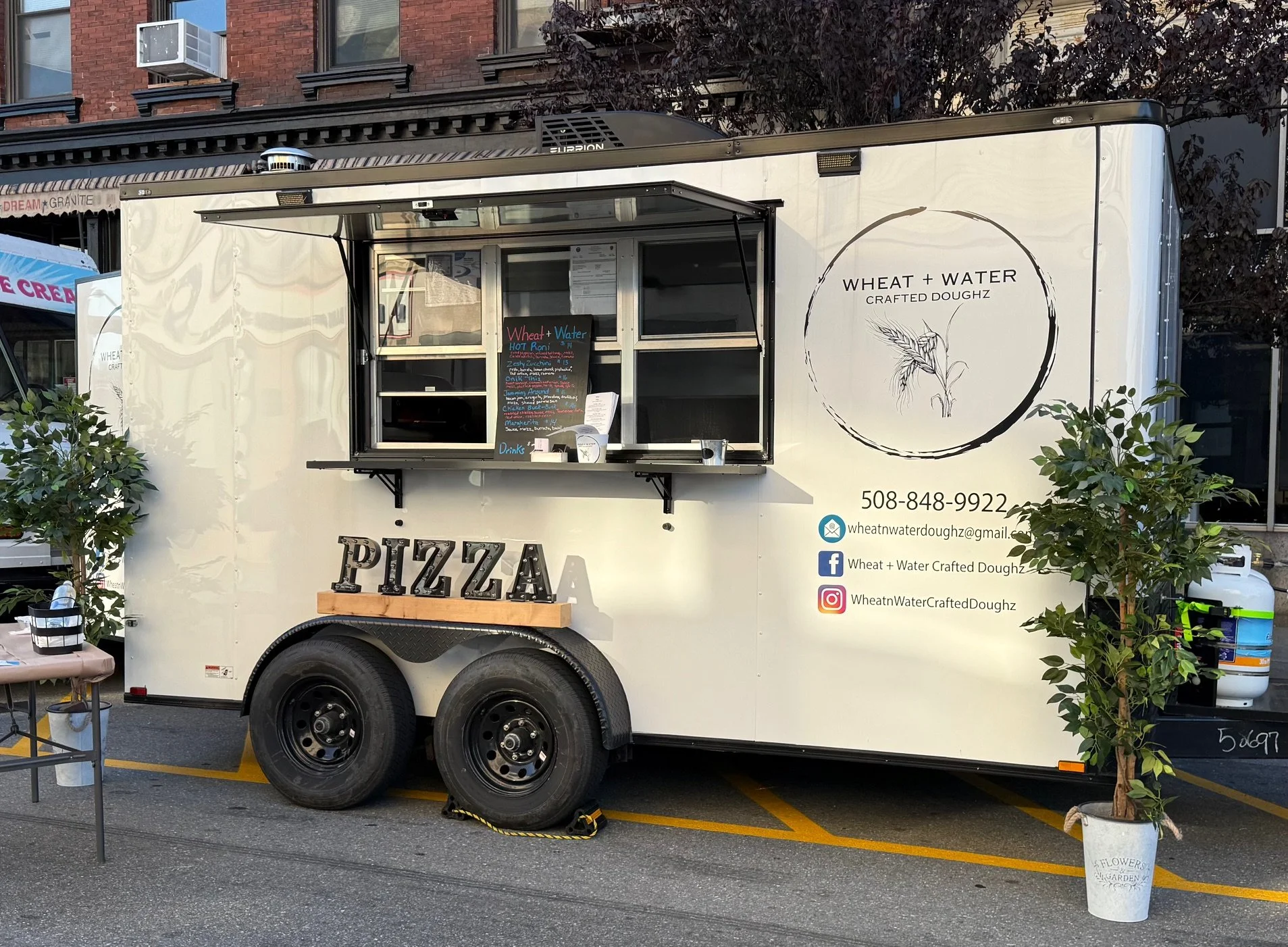 Food truck named Wheat + Water Crafted Doughz parked on street with green plants in white pots nearby, advertising pizza, with an menu board and social media icons on the side.