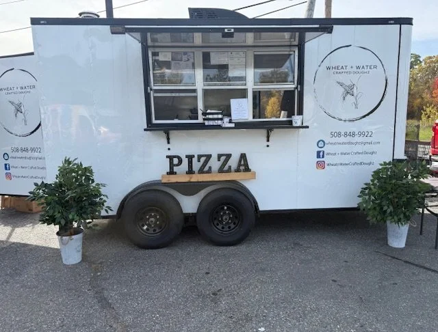 White food truck with a window, two black tires attached to the side, and a wooden sign spelling 'PIZZA' beneath the window. Two plants in white pots are positioned at the front, and the truck has logos and contact information for 'Wheat + Water' on both sides.