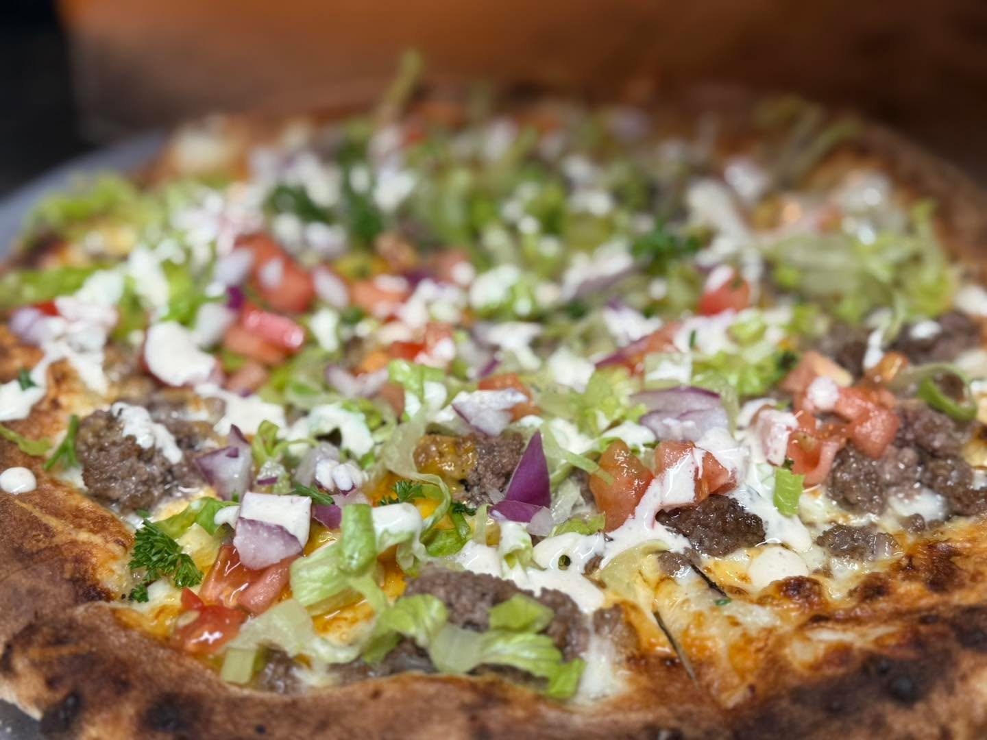 Close-up of a freshly baked pizza topped with ground beef, chopped tomatoes, diced red onions, shredded lettuce, and drizzled with white sauce.