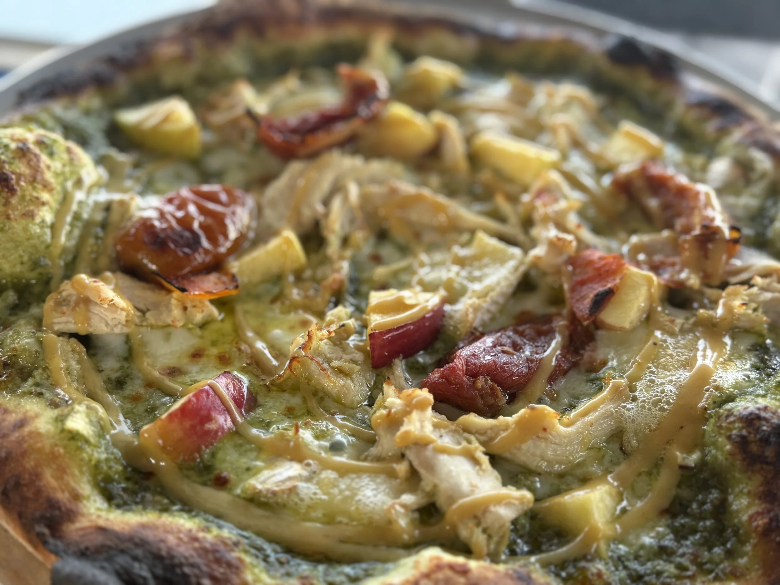 Close-up of a pizza with green pesto sauce, grilled chicken pieces, tomato slices, and melted cheese, with a slightly charred crust.