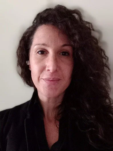 A woman with dark, curly hair and light skin poses for a portrait against a plain background.