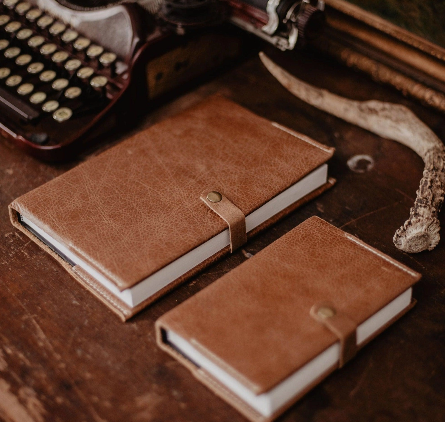 Brown leather-bound notebooks, an antique typewriter, and a long animal horn arranged on a wooden table.