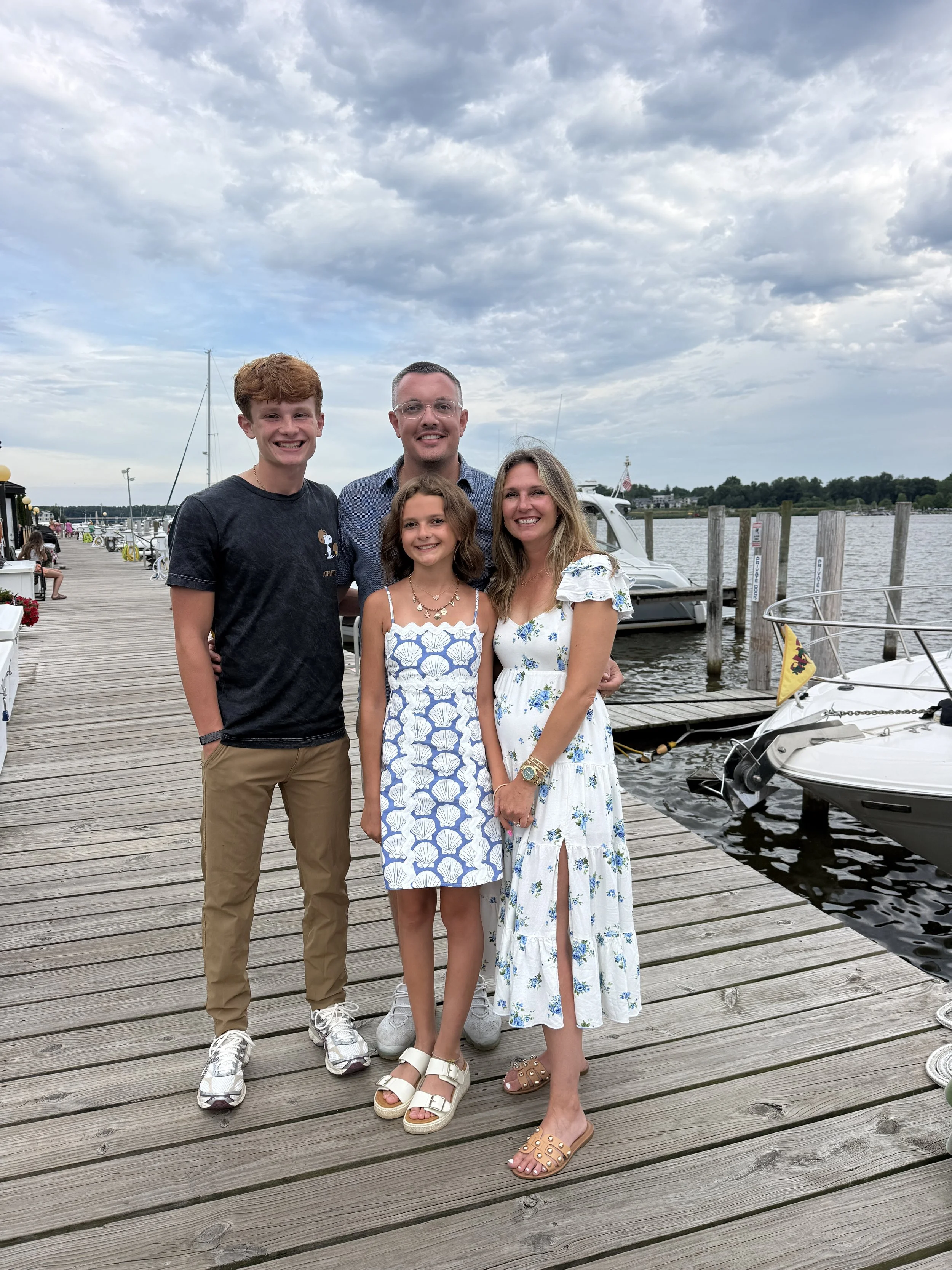 A family of four standing on a wooden dock near boats, with an overcast sky and water in the background.