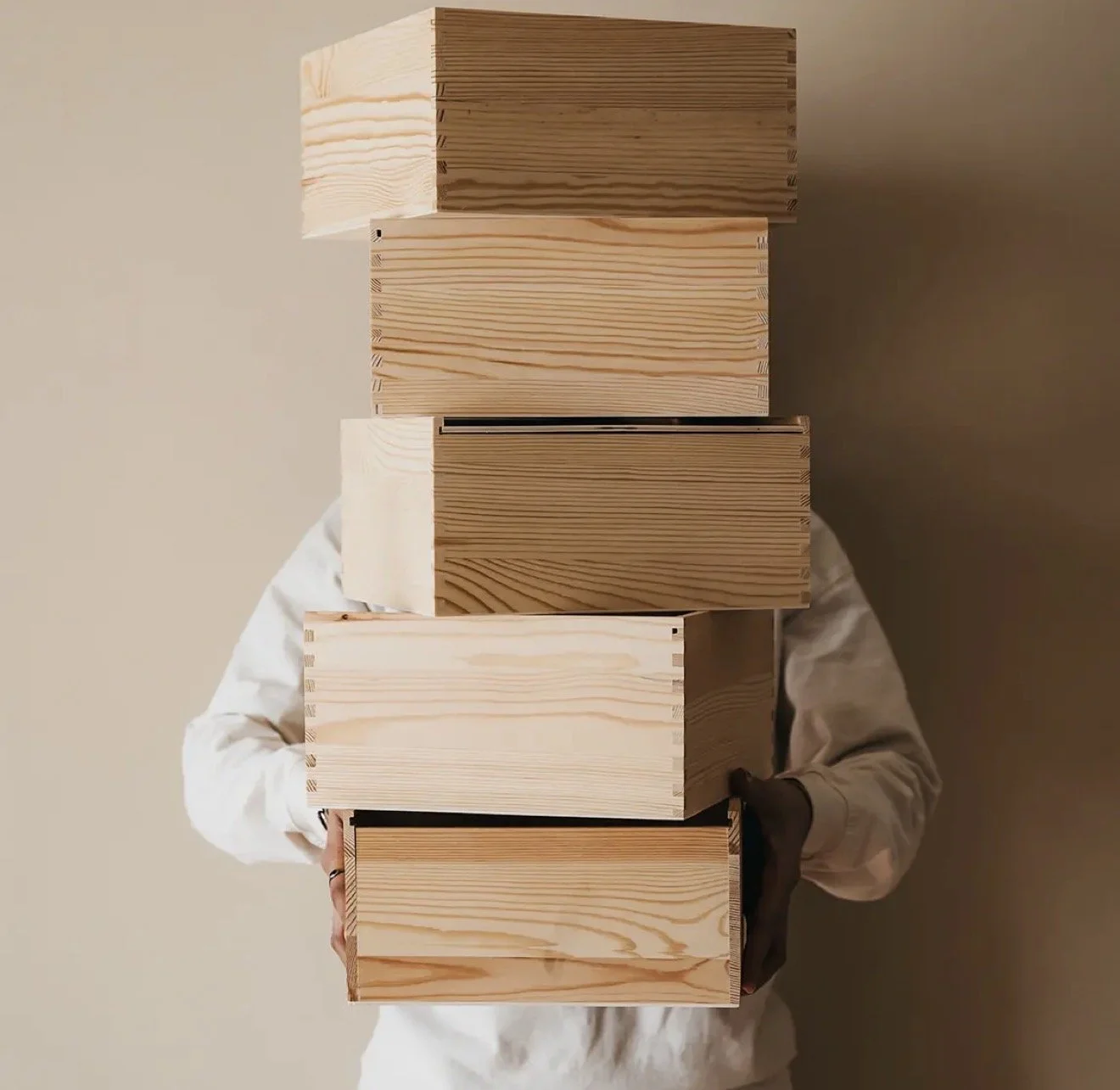 Person in white shirt holding five wooden boxes stacked vertically, obscuring their face, against a beige background.