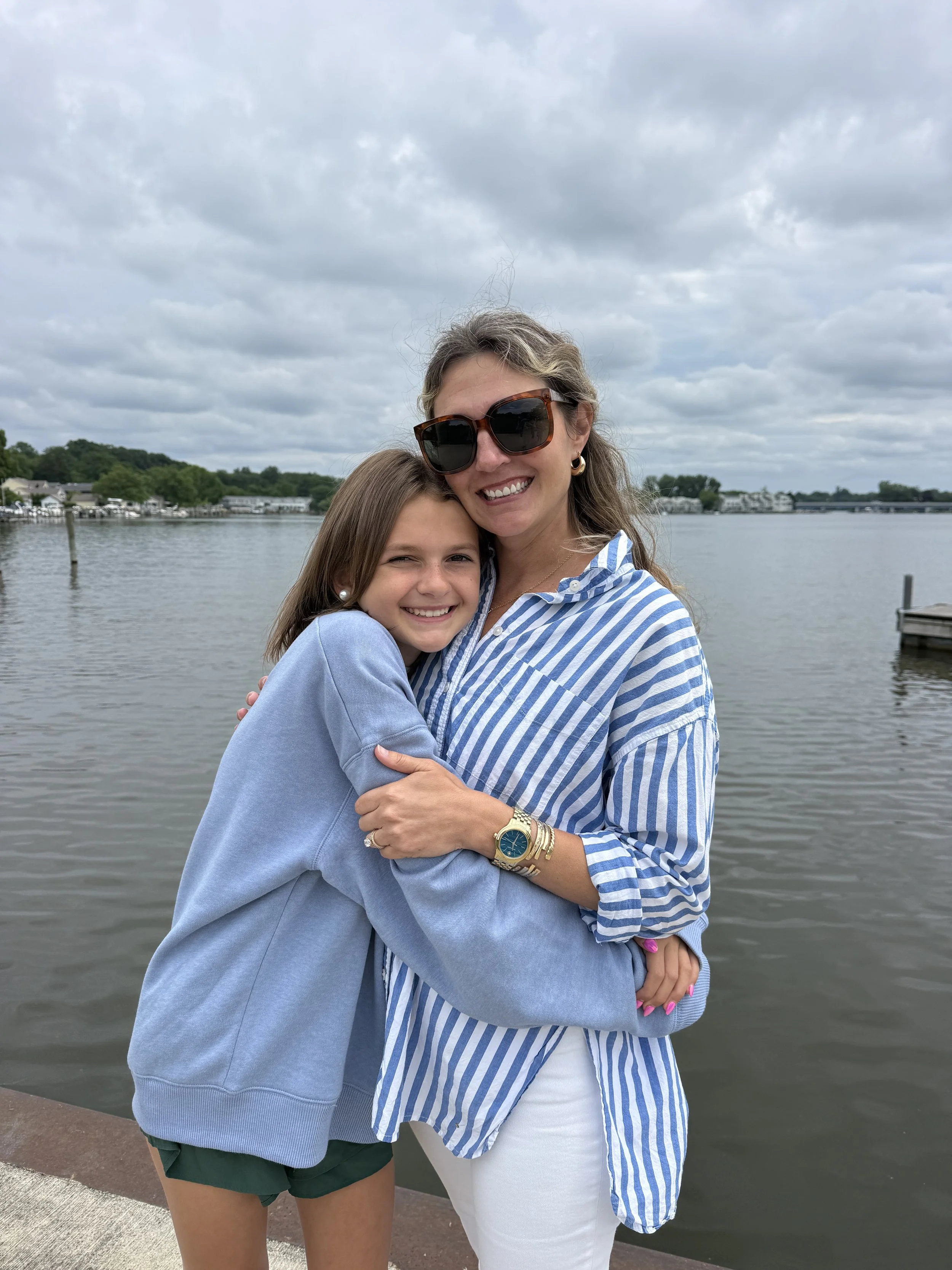 A woman and a girl embracing by a body of water outdoors, with a cloudy sky and houses visible in the background.
