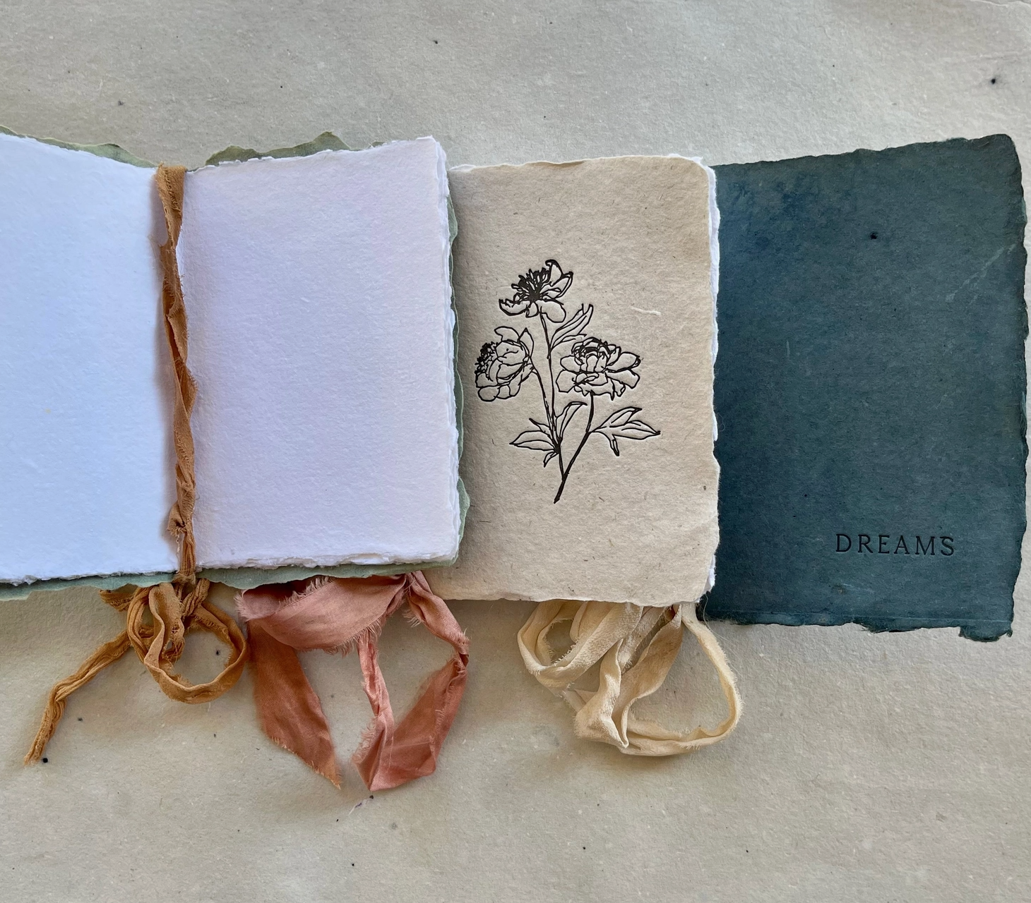 Three handmade paper pages connected with fabric ribbons, one featuring a line drawing of flowers and another with the word 'DREAMS' printed on a dark textured background.