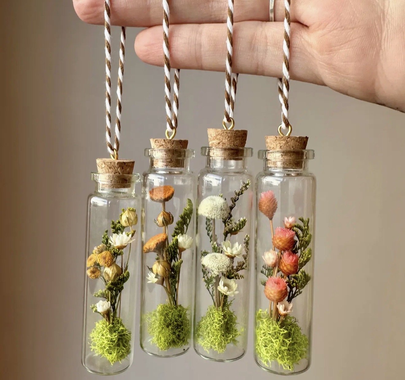 Three glass vials with cork stoppers hanging by twisted white and brown cords, each containing small dried flowers and green moss.