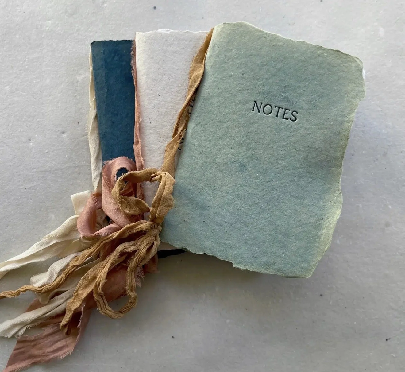 A bundle of handmade paper notebooks tied with pastel-colored fabric strips, with one notebook labeled 'NOTES' on a white textured surface.
