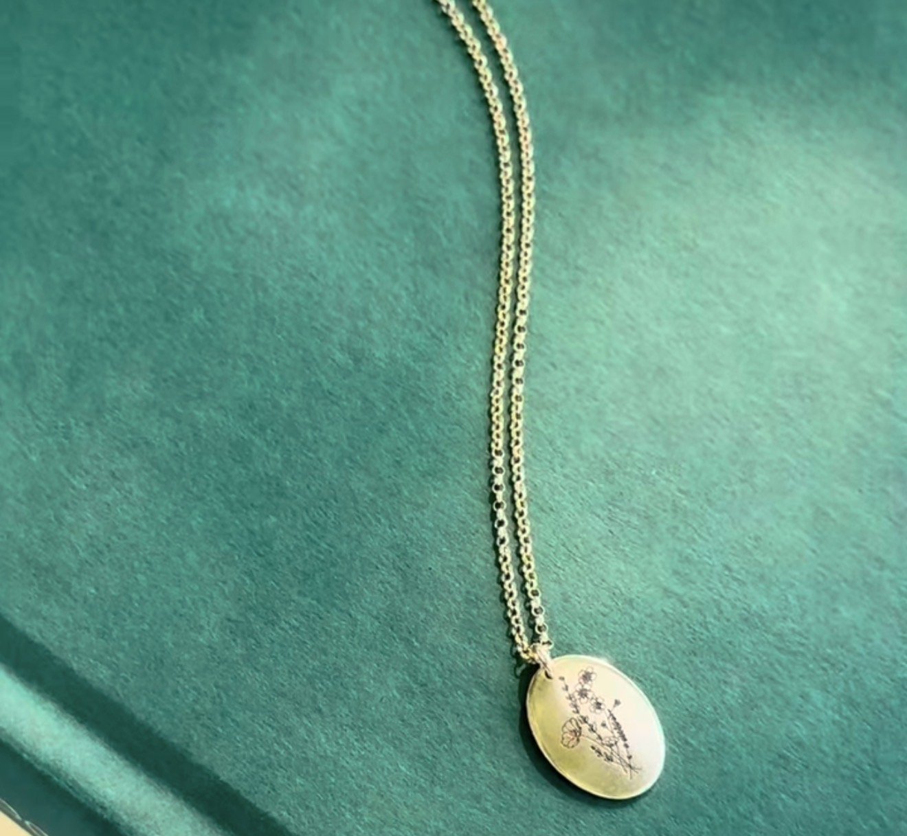 Gold necklace with an oval pendant engraved with a floral design, displayed on a green fabric background.