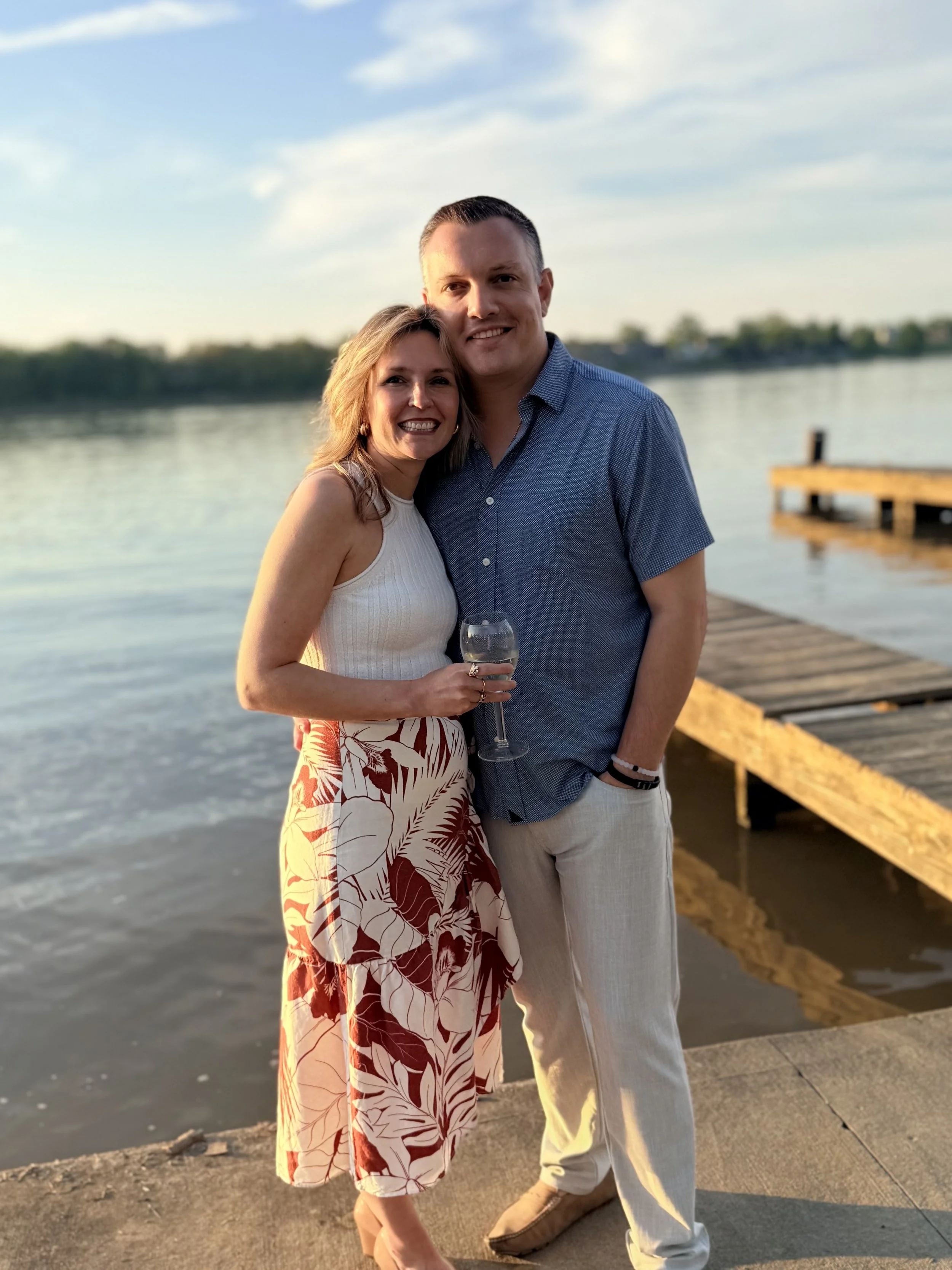 A smiling couple stands close together on a dock by a body of water during sunset, with the woman holding a glass of wine and wearing a white sleeveless top and a floral skirt, and the man wearing a blue short-sleeved shirt and white pants.