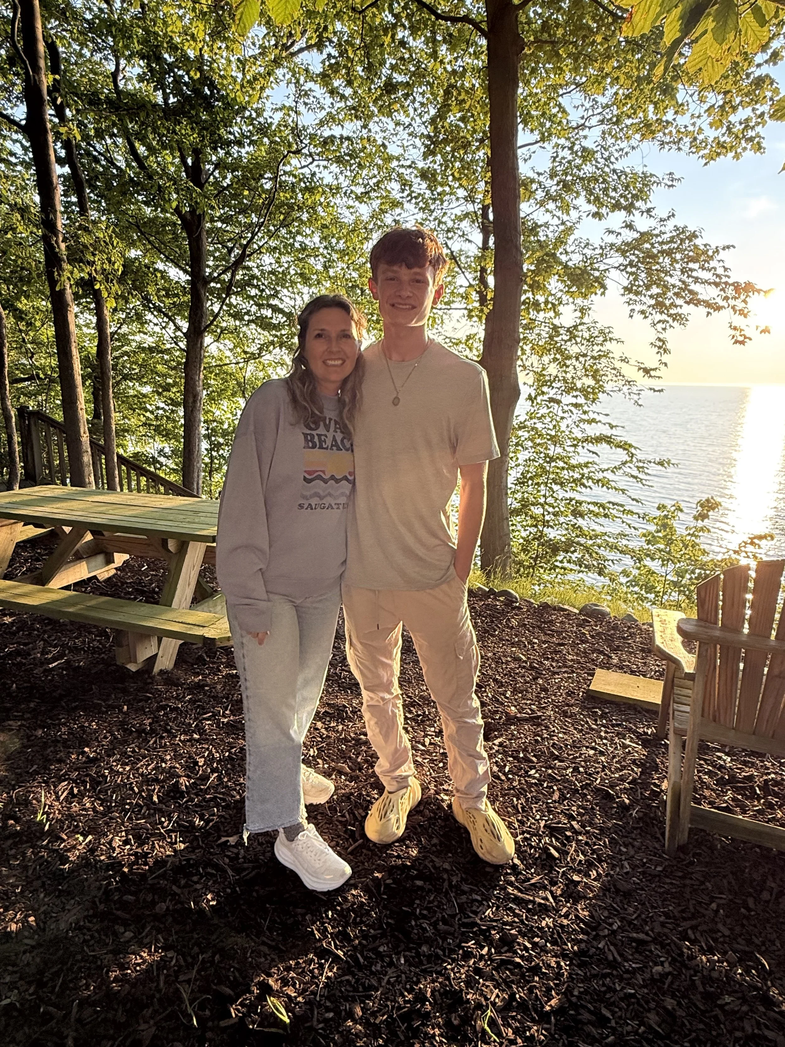 A woman and a young man standing outdoors near a lake with trees and benches in the background, during sunset.