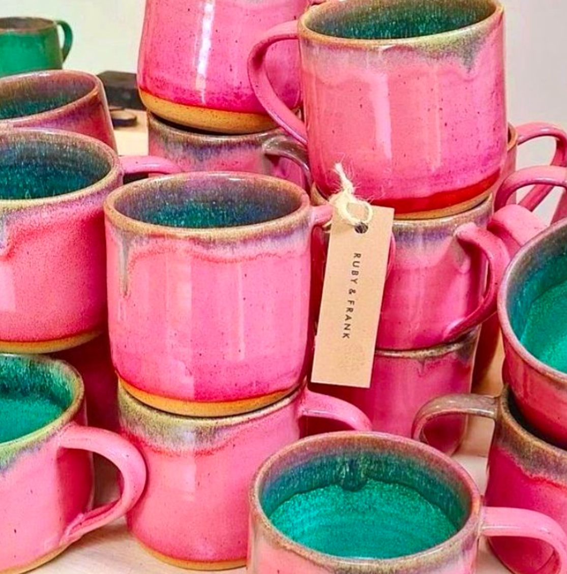A collection of colorful ceramic coffee mugs in pink, with some showing greenish-blue interiors, stacked or arranged together. The mugs are glazed with a vibrant, glossy finish.