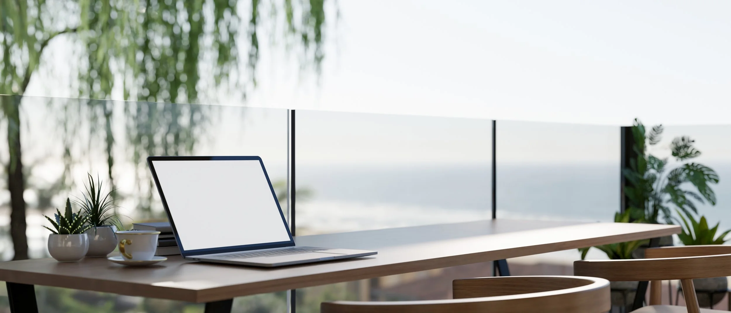 Empty desk with a laptop, two potted plants, a coffee cup, and a stack of papers, with a glass barrier and trees in the background.