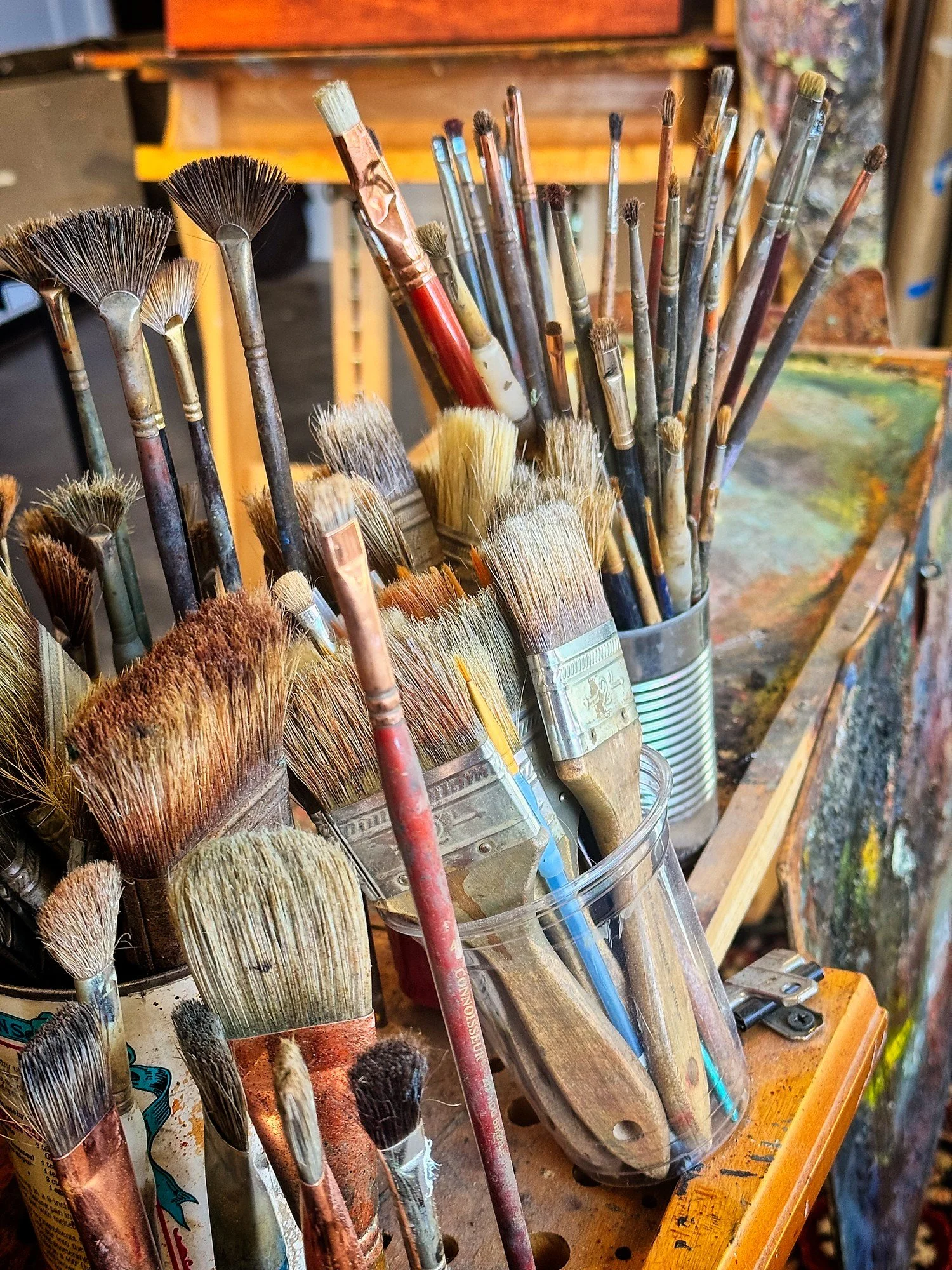 Various worn paintbrushes with different bristle types and sizes in a transparent container, on a painting workspace with a wooden table and background