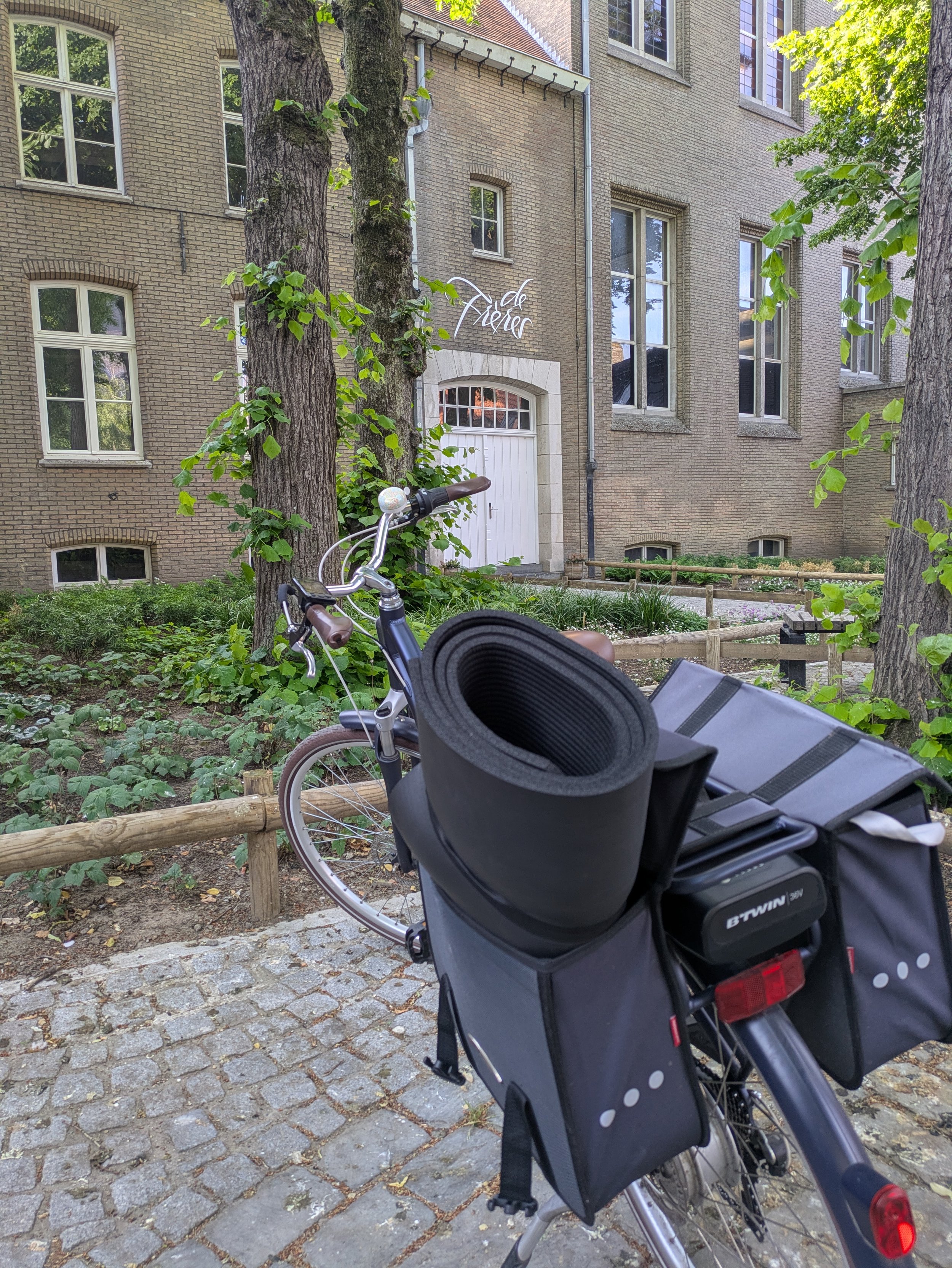 A bicycle parked on a cobblestone sidewalk in front of a brick building with large windows and a white door. There are two large trees and green foliage. A yoga mat and bag are attached to the bike's back.