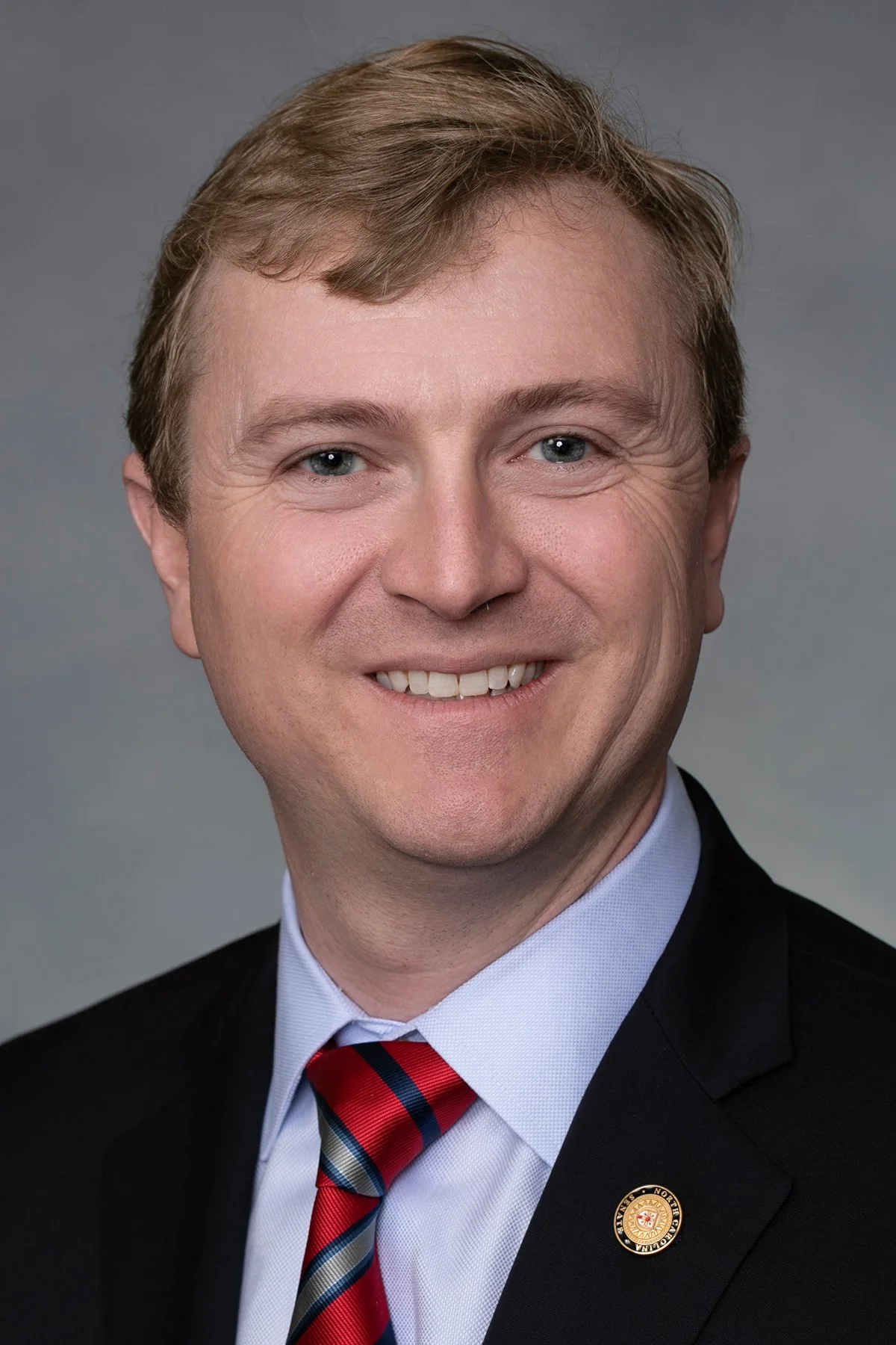 A middle-aged man with light brown hair, blue eyes, and fair skin, smiling, wearing a black suit, a white dress shirt, a red and blue striped tie, and a pin on his lapel, against a gray background.