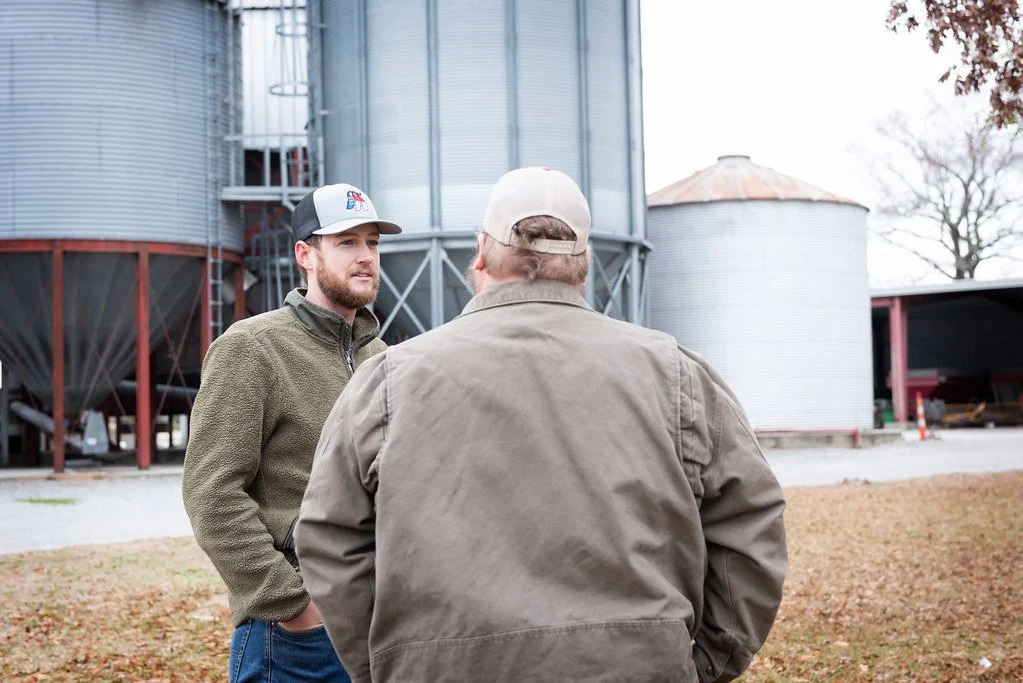 Two men talking outdoors in front of storage silos on a farm, one wearing a baseball cap and the other with a backwards cap.