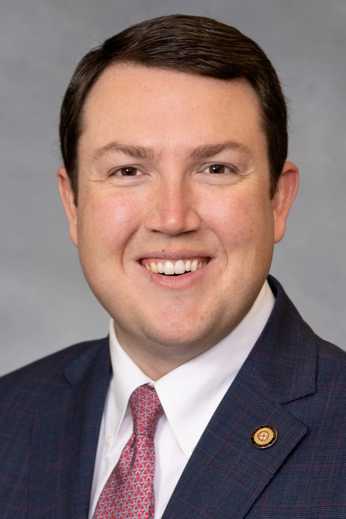 Headshot of a man with brown hair, smiling, wearing a dark checkered suit, white shirt, and a patterned red tie, featuring a lapel pin.