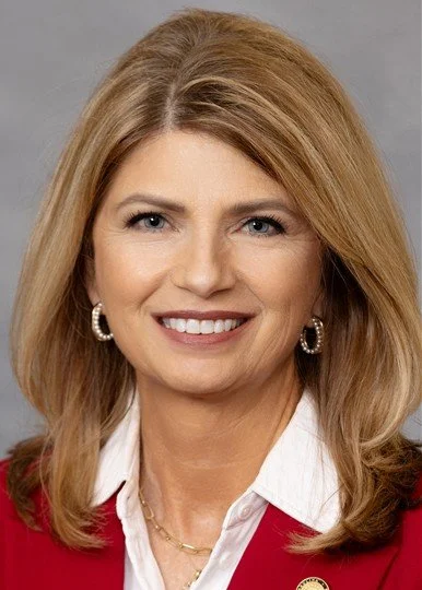 Portrait of a smiling woman with shoulder-length blonde hair, wearing pearl earrings and a red blazer over a white shirt.