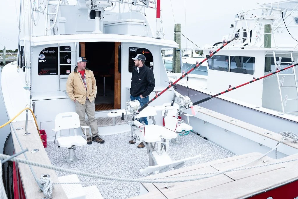 Two men standing on a fishing boat deck, engaged in conversation, with fishing rods secured on the right side and other boats docked nearby.