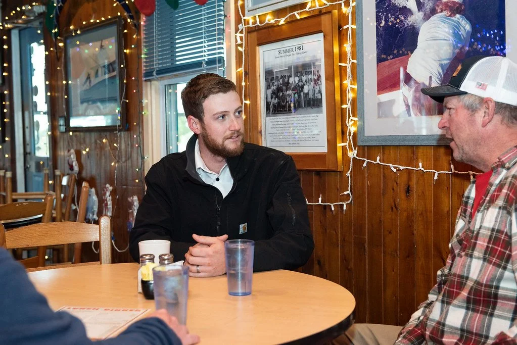 Two men sitting at a wooden table in a cozy restaurant decorated with string lights and framed photos, engaged in conversation.