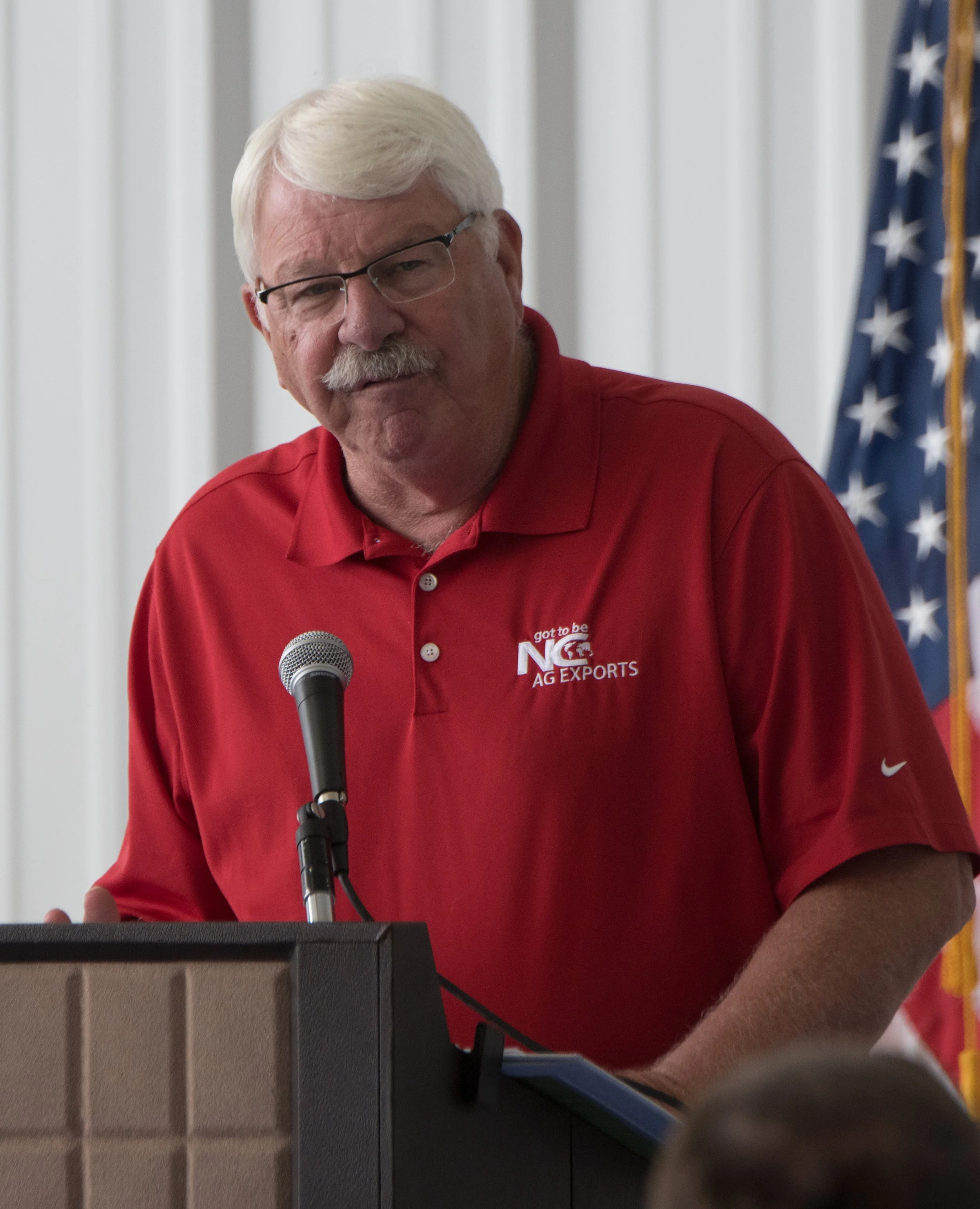 A man with white hair and glasses wearing a red polo shirt with 'Got to be N C AG EXPORTS' logo, standing at a podium with a microphone, with an American flag in the background.